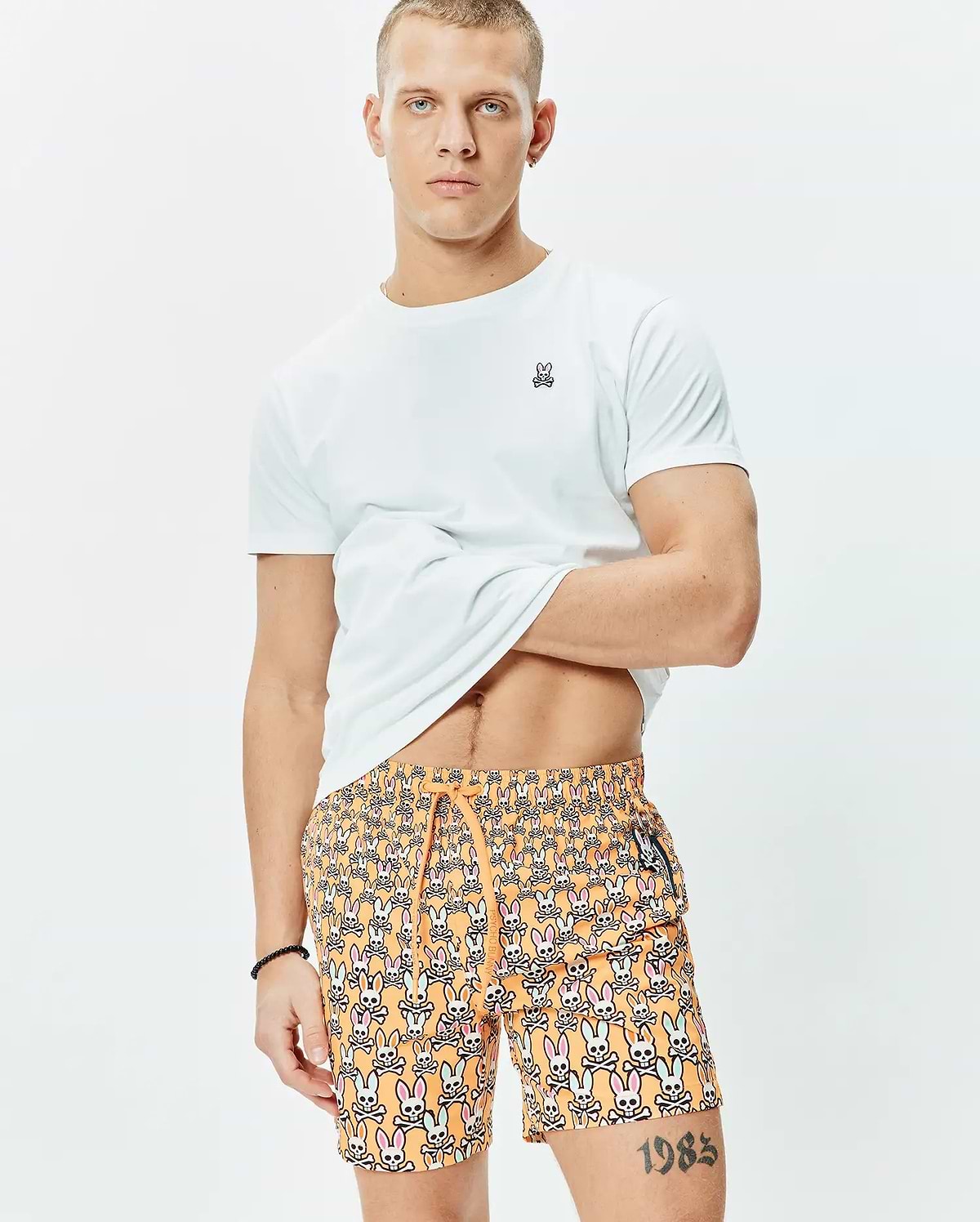 White classic fit jersey t-shirt with small embroidered Psycho Bunny logo on left chest, paired with orange Mens Byron Swim Trunk 6 featuring an all-over multi-color bunny skull print and drawstring waist.