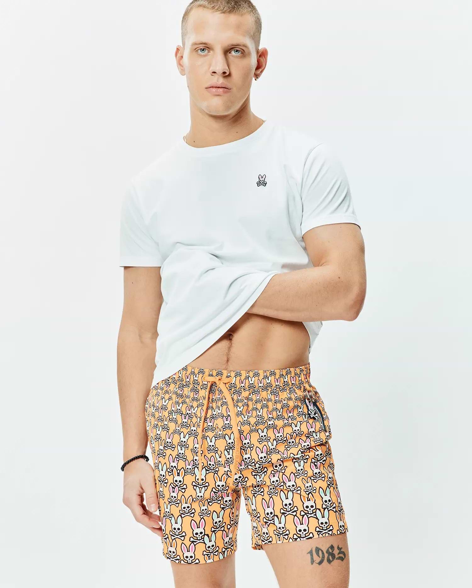 White classic fit jersey t-shirt with small embroidered Psycho Bunny logo on left chest, paired with orange Mens Byron Swim Trunk 6 featuring an all-over multi-color bunny skull print and drawstring waist.