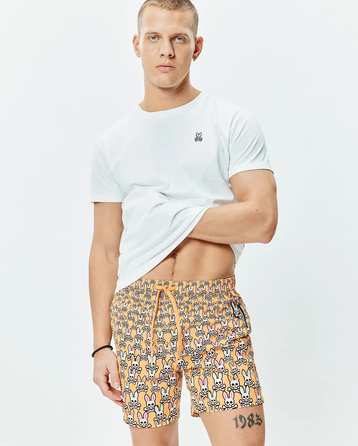 White classic fit jersey t-shirt with small embroidered Psycho Bunny logo on left chest, paired with orange Mens Byron Swim Trunk 6 featuring an all-over multi-color bunny skull print and drawstring waist.