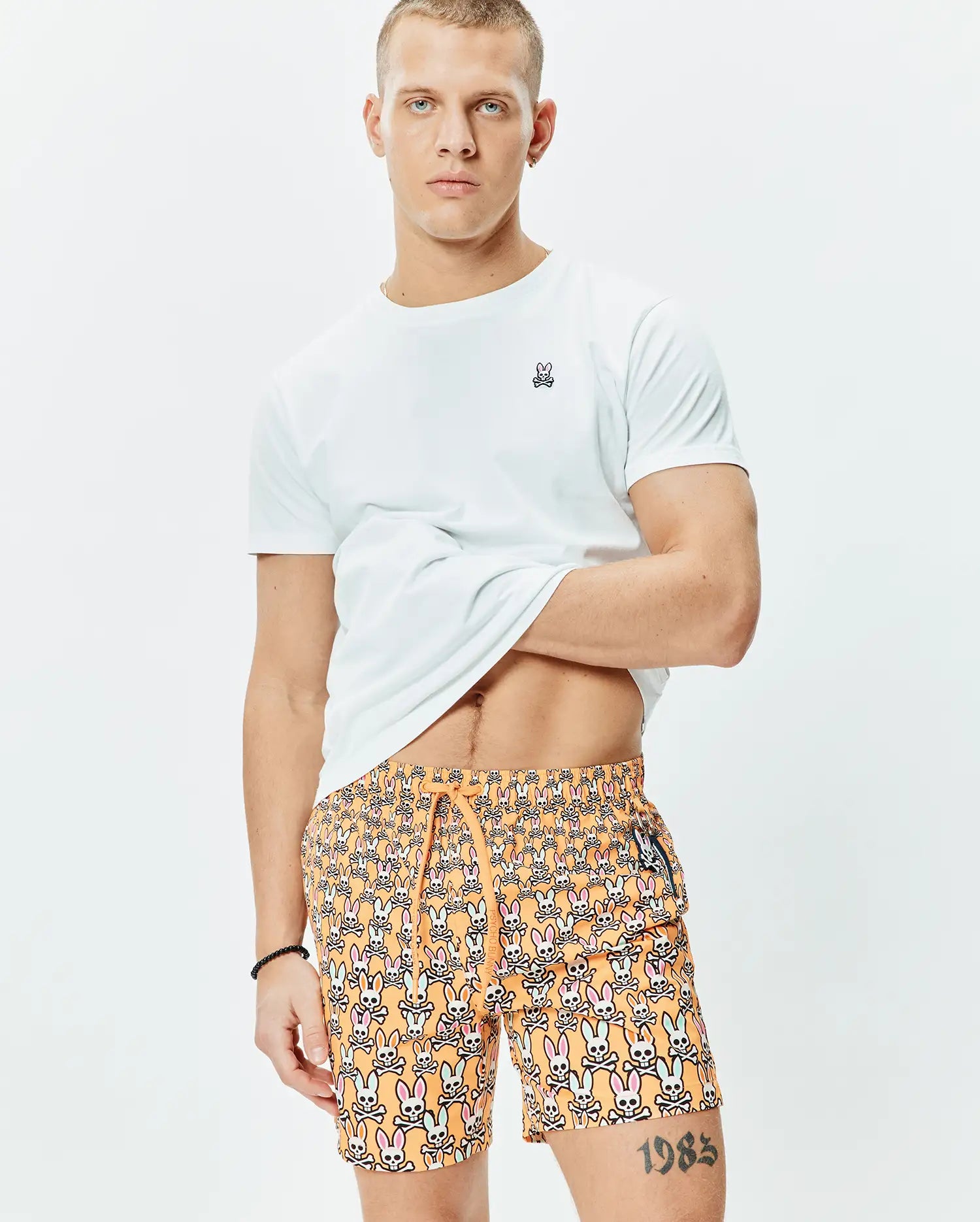 White classic fit jersey t-shirt with small embroidered Psycho Bunny logo on left chest, paired with orange Mens Byron Swim Trunk 6 featuring an all-over multi-color bunny skull print and drawstring waist.