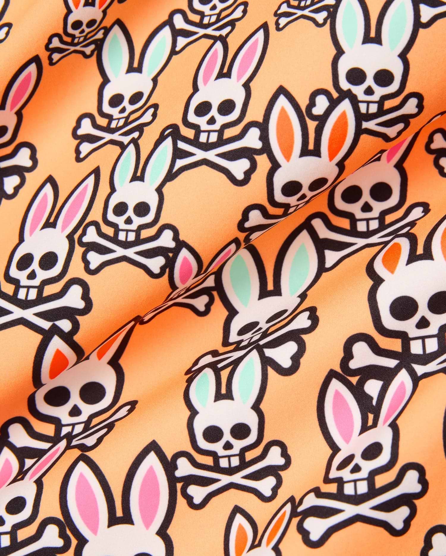 Mens Byron Swim Trunk 6, peach with an all-over print of the iconic Psycho Bunny skull logo, featuring white skulls with black outlines and alternating pink, mint green, and orange ear accents on smooth swim fabric.