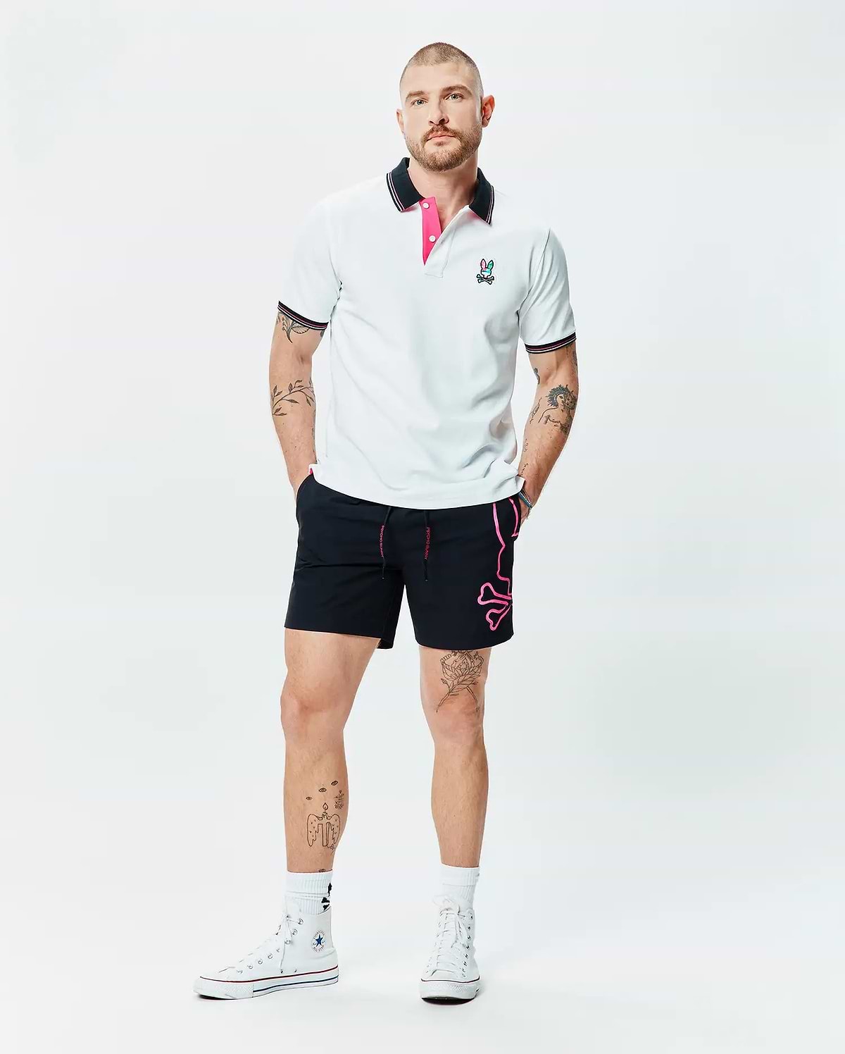White pique polo with black and pink striped collar, embroidered Psycho Bunny logo, and black Mens Duncan Bunny Print Swim Trunk featuring a large pink bunny skull graphic and pink drawstring.