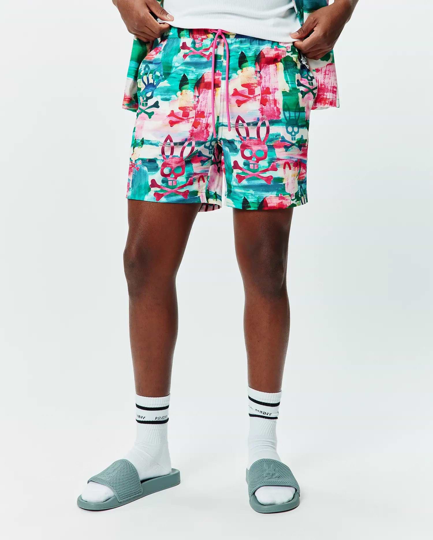 Mens Nate All Over Print Swim Trunk in a vibrant multi-color abstract brushstroke pattern featuring repeated Psycho Bunny skull and crossbones logos, with a pink drawstring, offering a relaxed fit.
