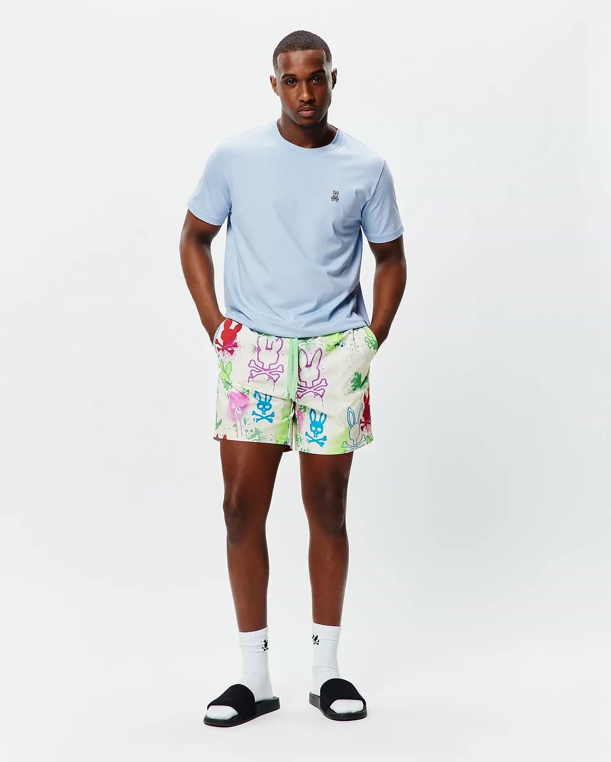 Light blue classic fit jersey t-shirt with small embroidered Psycho Bunny logo on left chest. Mens Quintin All Over Print Swim Trunk features an off-white base, vibrant multi-color graffiti-style bunny skull print, and neon green drawstring.