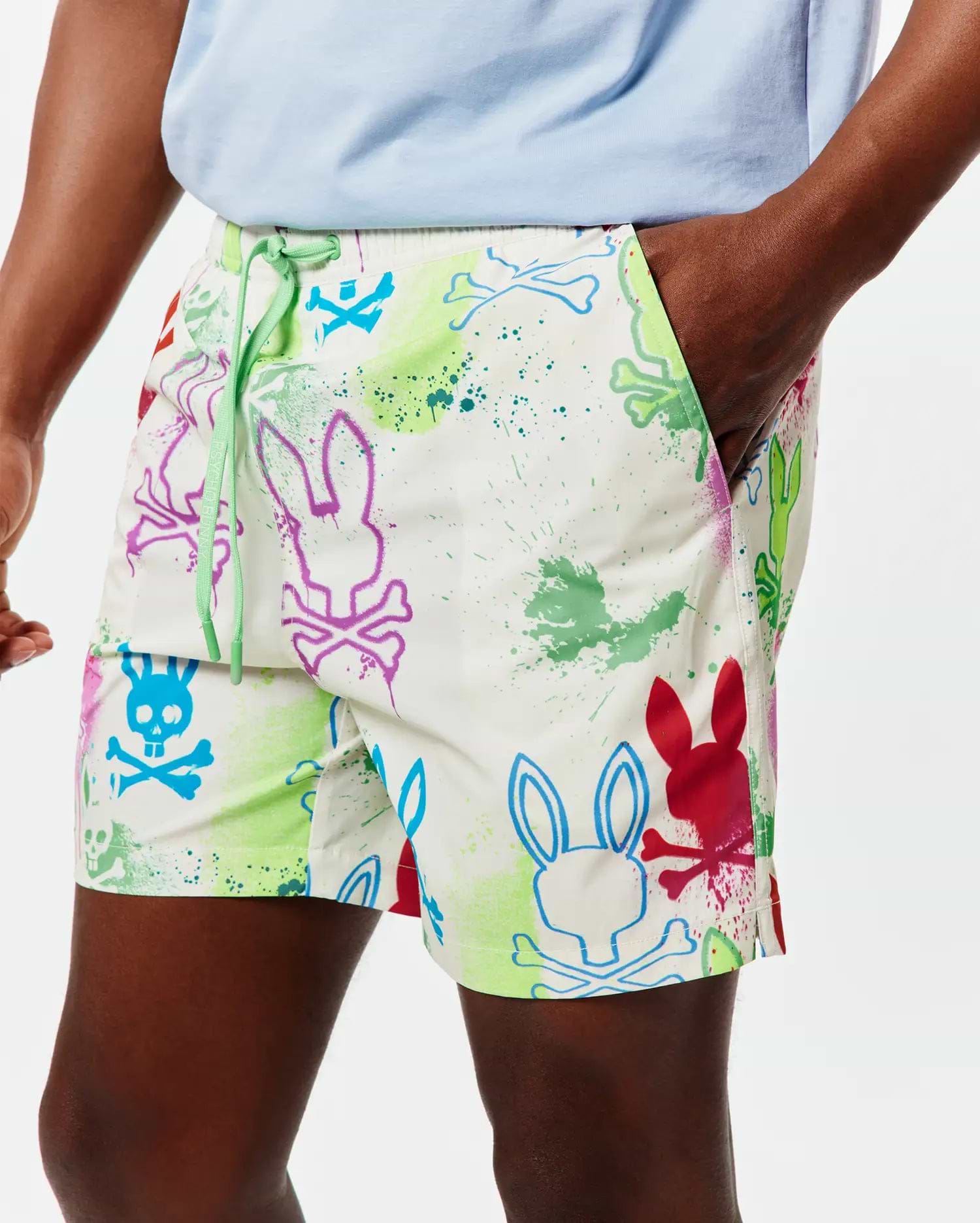 Mens Quintin All Over Print Swim Trunk in white, with a vibrant multicolor paint splatter and outlined Psycho Bunny skull logo pattern, featuring a relaxed fit and green drawstring with PSYCHO BUNNY text.