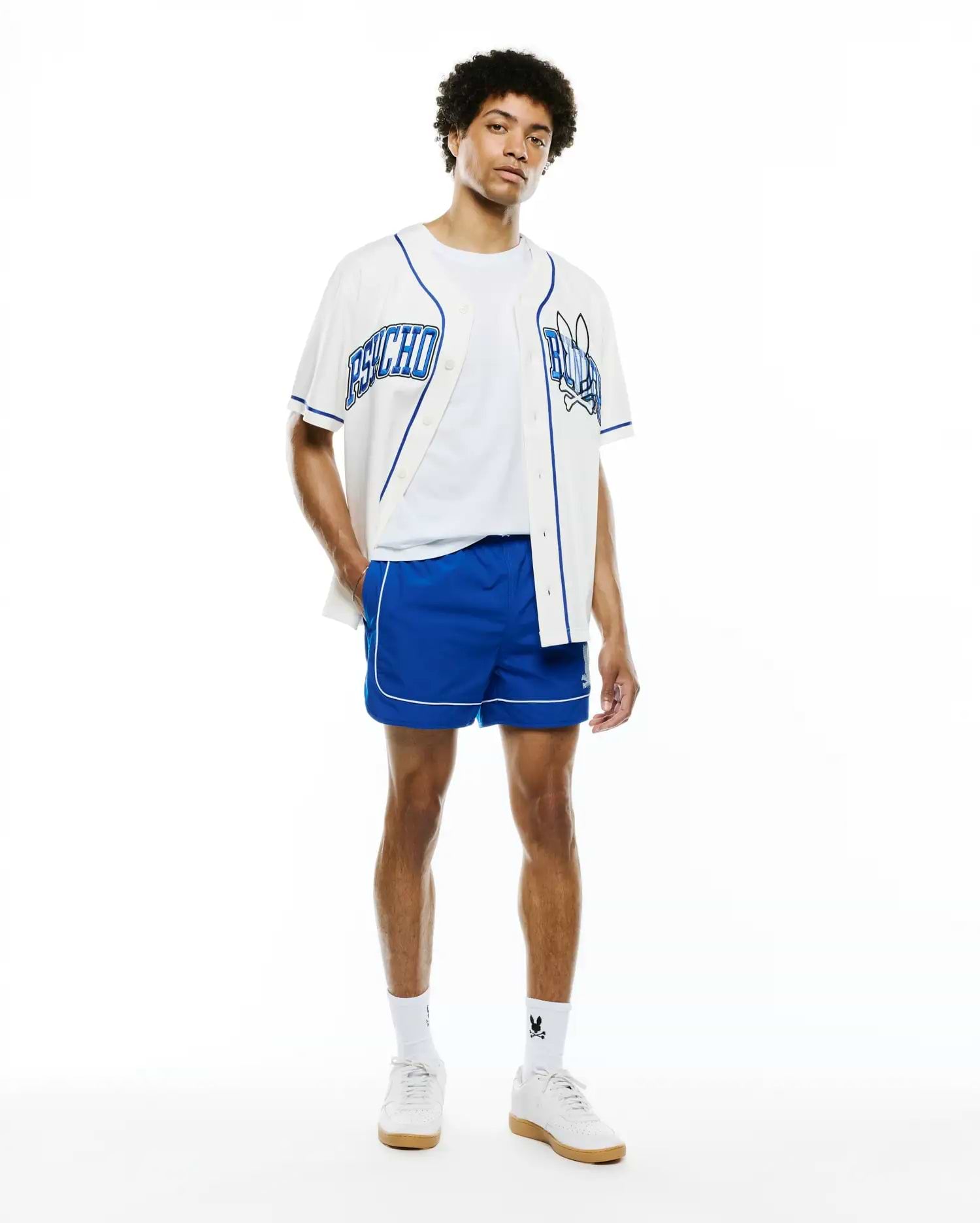 Mens Nathaniel Swim Trunk in royal blue with white piping, paired with a white baseball jersey featuring blue Psycho text and skull logo, over a white tee, and white sneakers with Psycho Bunny socks.