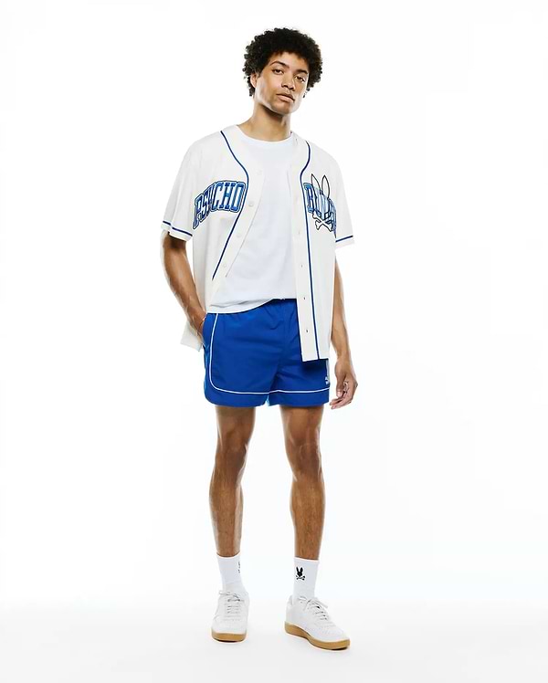 Mens Nathaniel Swim Trunk in royal blue with white piping, paired with a white baseball jersey featuring blue Psycho text and skull logo, over a white tee, and white sneakers with Psycho Bunny socks.