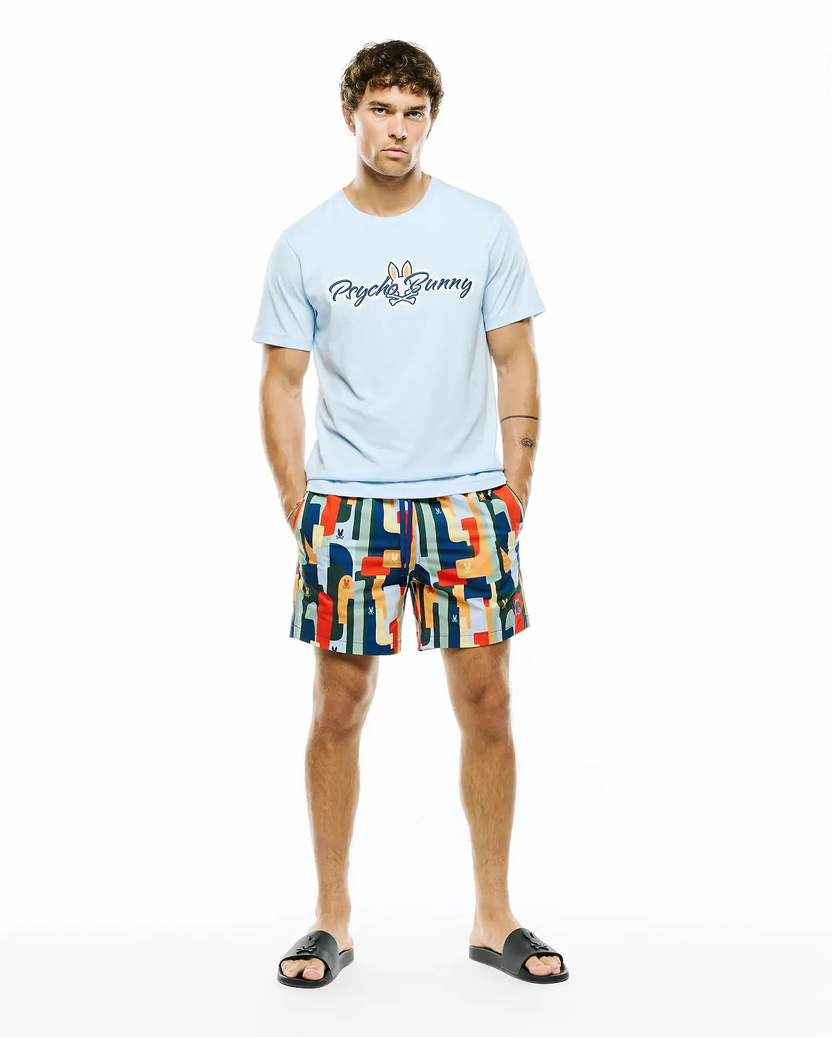 Light blue jersey t-shirt with large Psycho Bunny script logo on chest, paired with Mens Wes All Over Print Swim Trunk featuring a vibrant multi-color abstract pattern and subtle bunny skull logos.