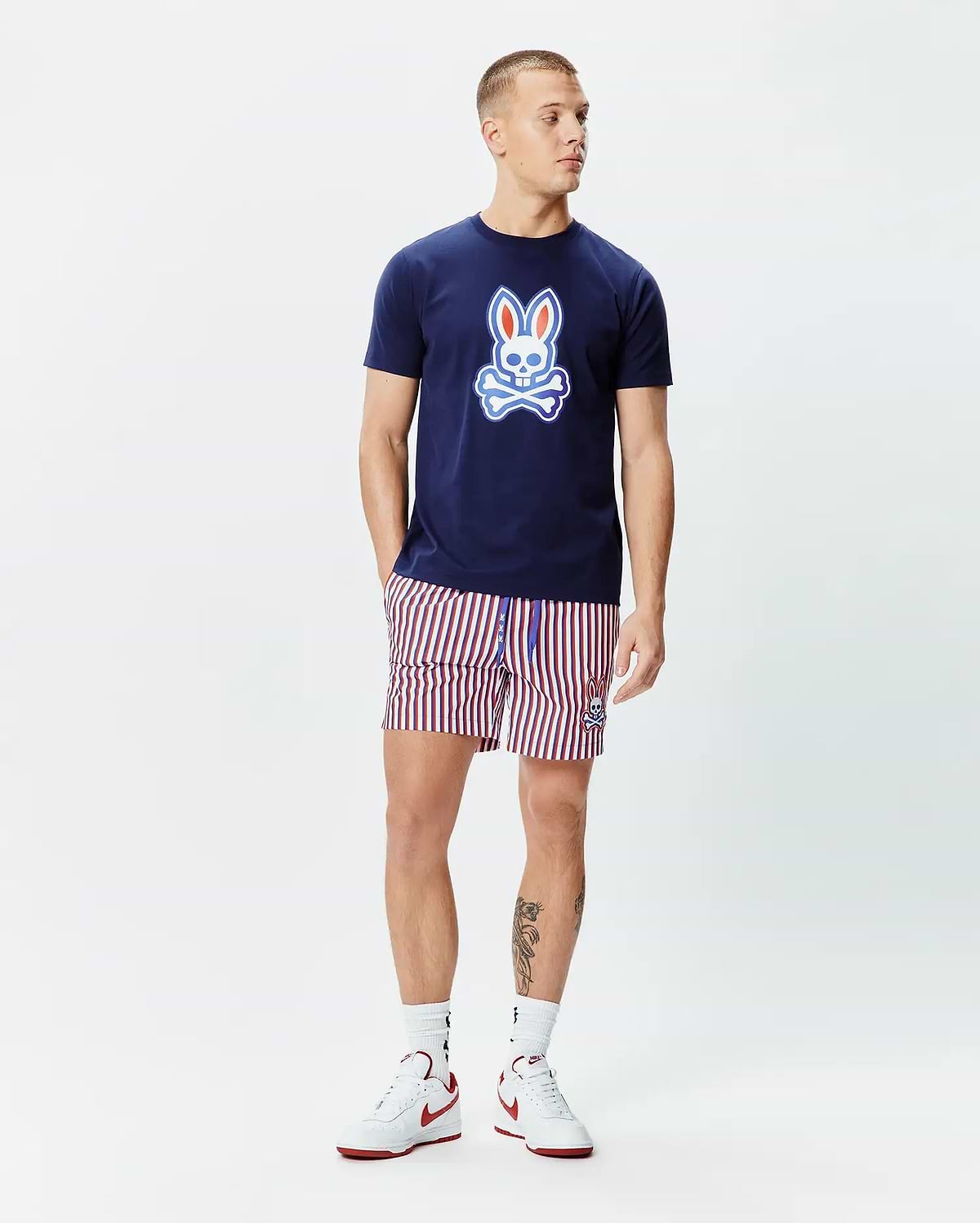 Navy blue jersey t-shirt with a large gradient Psycho Bunny skull logo on the chest, paired with Mens John All Over Print Swim Trunk featuring red, white, and navy vertical stripes and a small embroidered bunny logo.