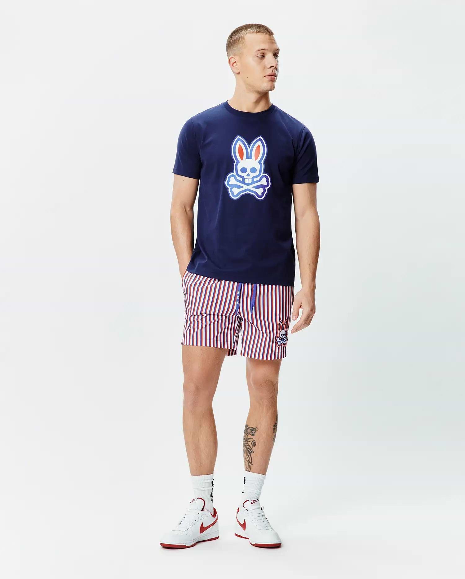 Navy blue jersey t-shirt with a large gradient Psycho Bunny skull logo on the chest, paired with Mens John All Over Print Swim Trunk featuring red, white, and navy vertical stripes and a small embroidered bunny logo.