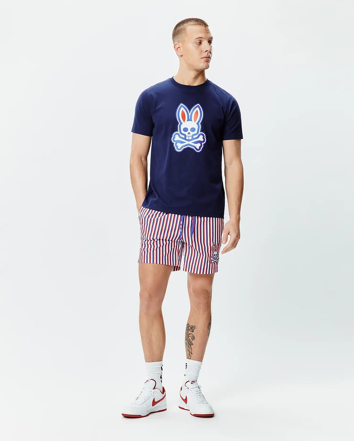 Navy blue jersey t-shirt with a large gradient Psycho Bunny skull logo on the chest, paired with Mens John All Over Print Swim Trunk featuring red, white, and navy vertical stripes and a small embroidered bunny logo.