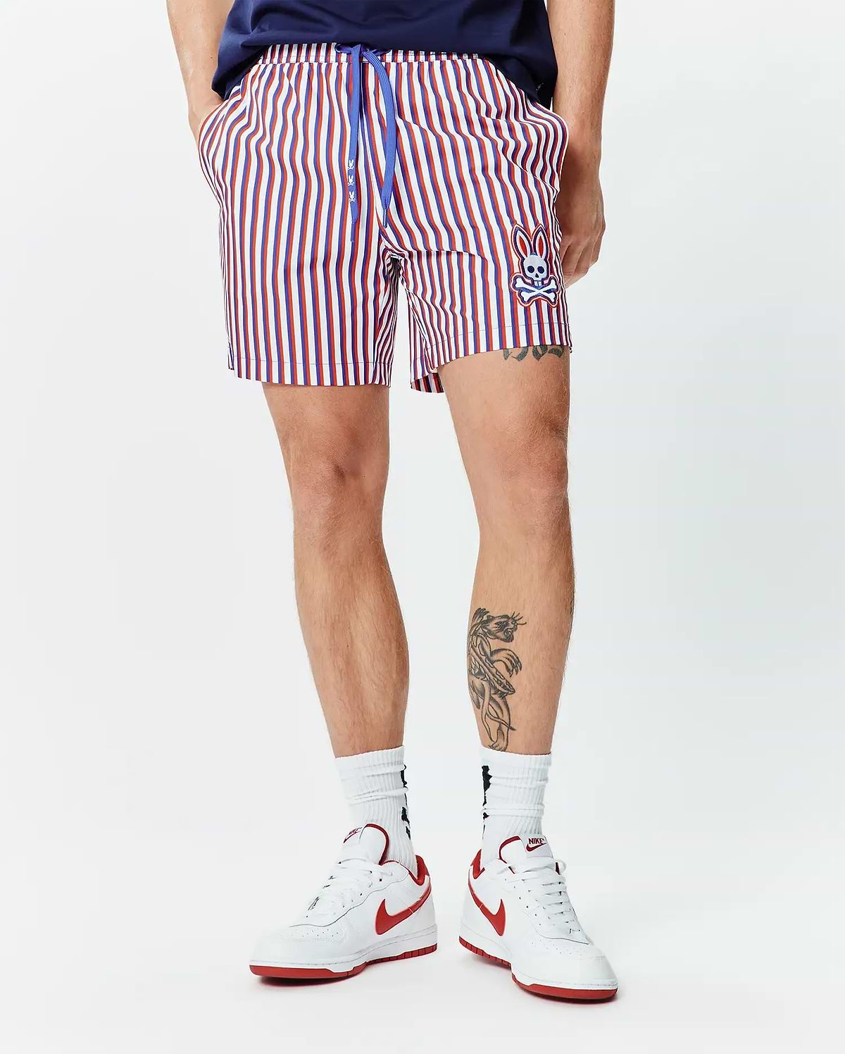 Mens John All Over Print Swim Trunk. Classic fit, red, white, and blue striped swim trunk with a blue drawstring featuring small bunny logos and an embroidered Psycho Bunny skull on the lower left leg.