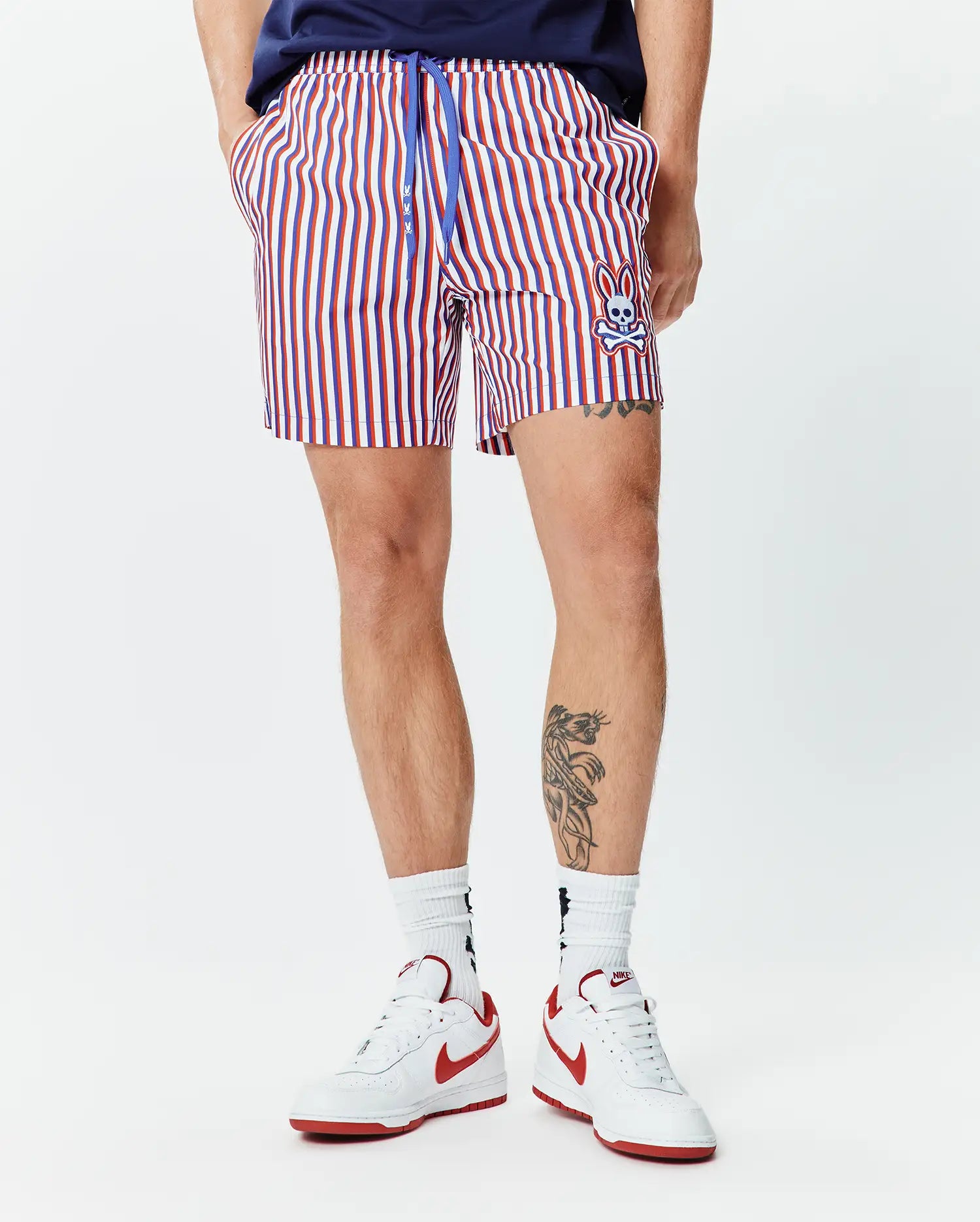 Mens John All Over Print Swim Trunk. Classic fit, red, white, and blue striped swim trunk with a blue drawstring featuring small bunny logos and an embroidered Psycho Bunny skull on the lower left leg.