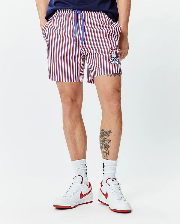 Mens John All Over Print Swim Trunk. Classic fit, red, white, and blue striped swim trunk with a blue drawstring featuring small bunny logos and an embroidered Psycho Bunny skull on the lower left leg.