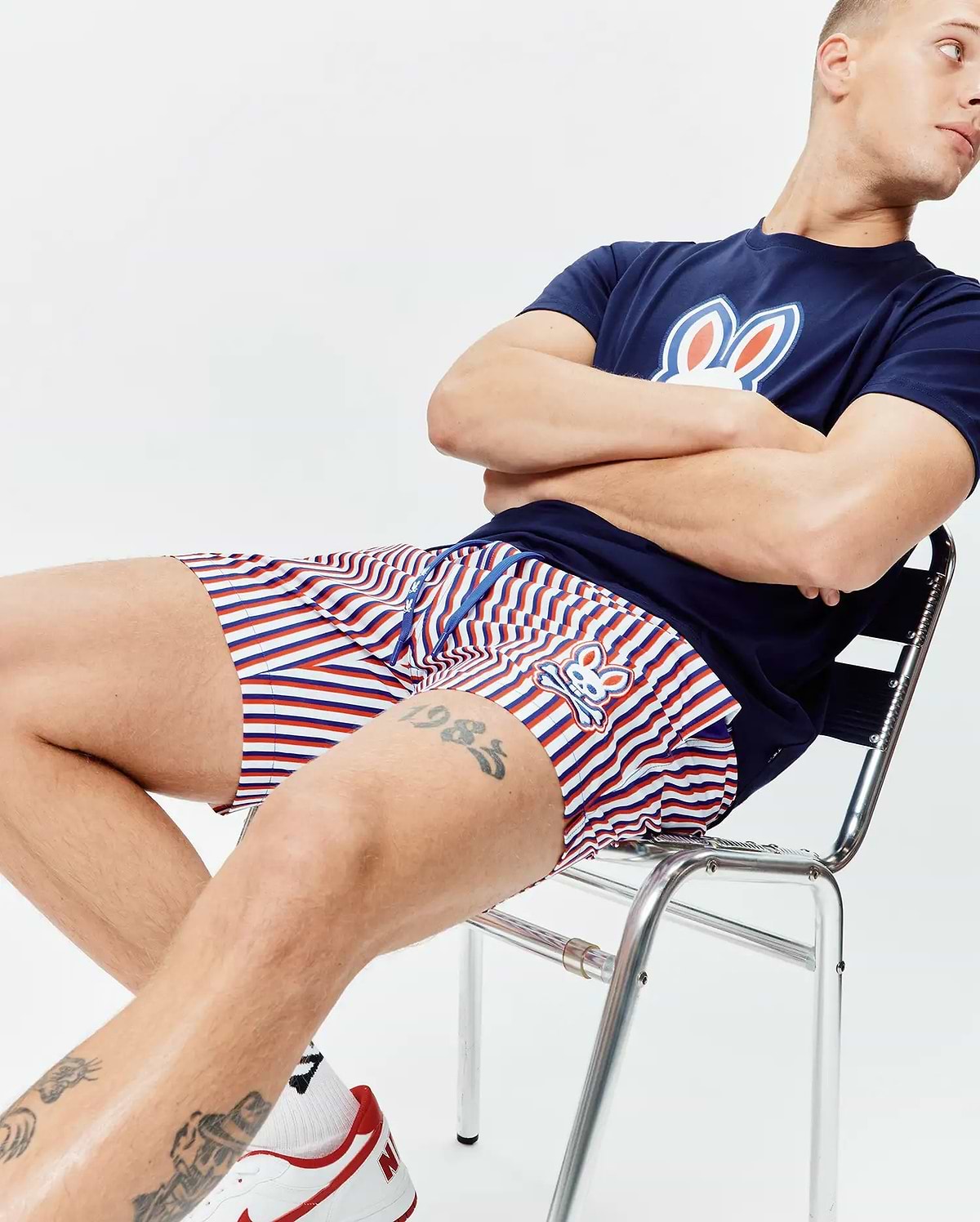 Navy blue jersey t-shirt with a large white, red, and blue Psycho Bunny logo on the chest, paired with Mens John All Over Print Swim Trunk featuring red, white, and blue stripes and a small embroidered bunny logo.