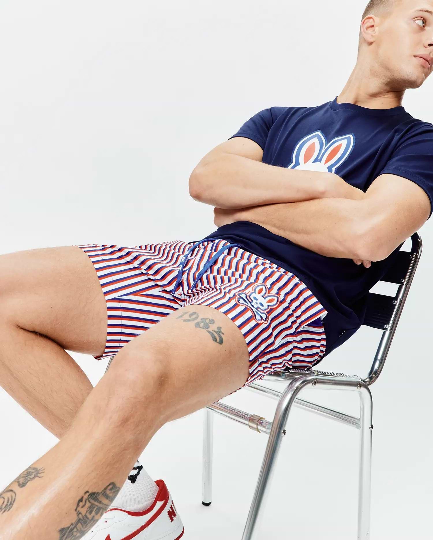Navy blue jersey t-shirt with a large white, red, and blue Psycho Bunny logo on the chest, paired with Mens John All Over Print Swim Trunk featuring red, white, and blue stripes and a small embroidered bunny logo.