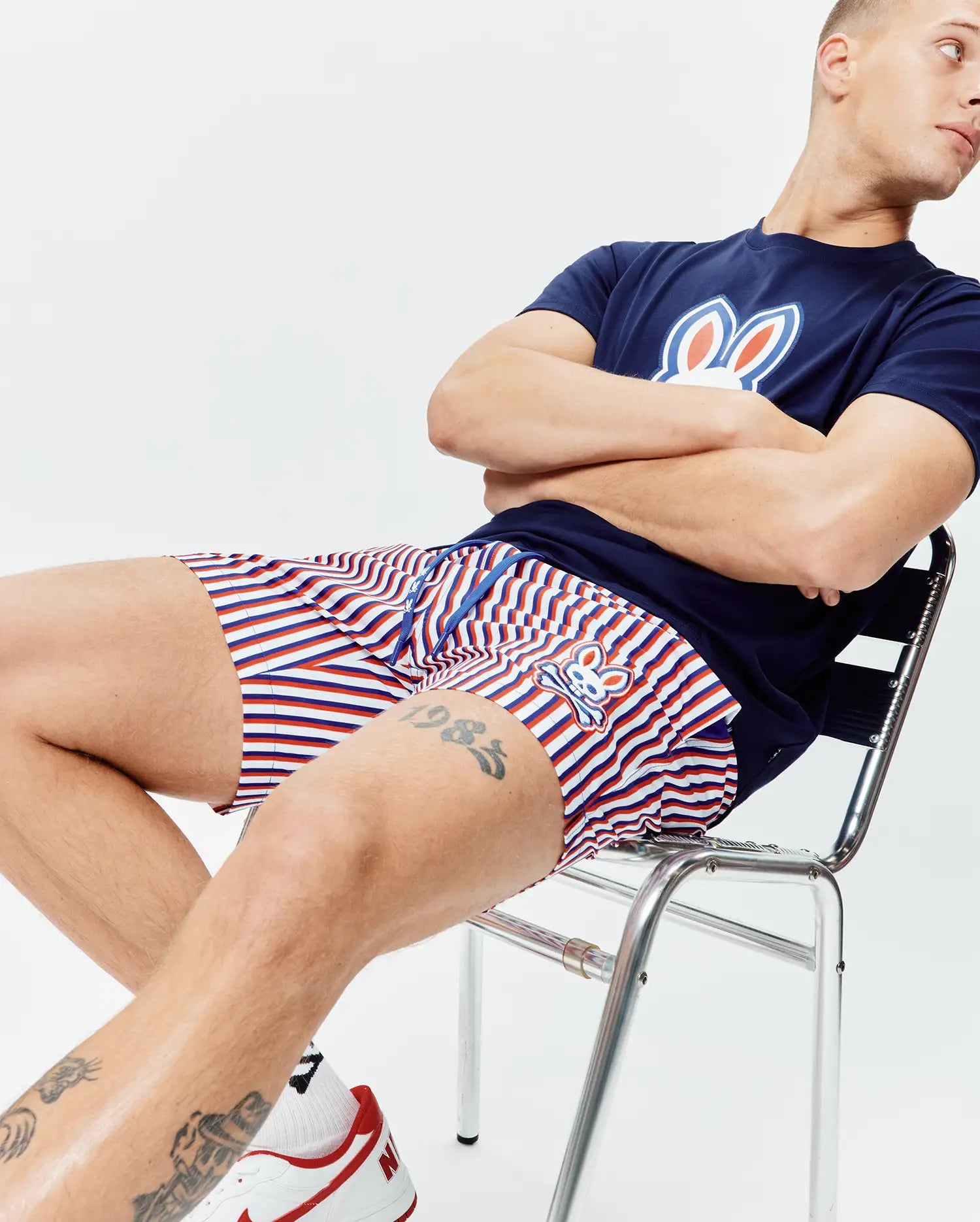 Navy blue jersey t-shirt with a large white, red, and blue Psycho Bunny logo on the chest, paired with Mens John All Over Print Swim Trunk featuring red, white, and blue stripes and a small embroidered bunny logo.