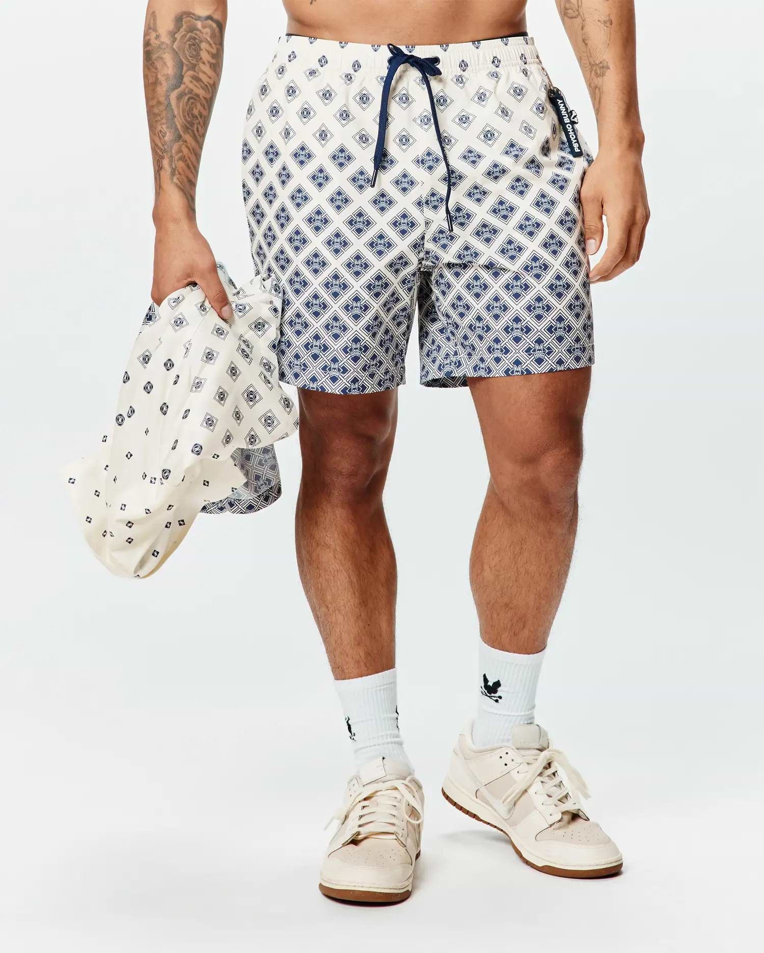 Mens Tobias Swim Trunk in cream with a navy geometric diamond pattern featuring integrated Psycho Bunny skull logos, a navy drawstring, and a branded side tag.