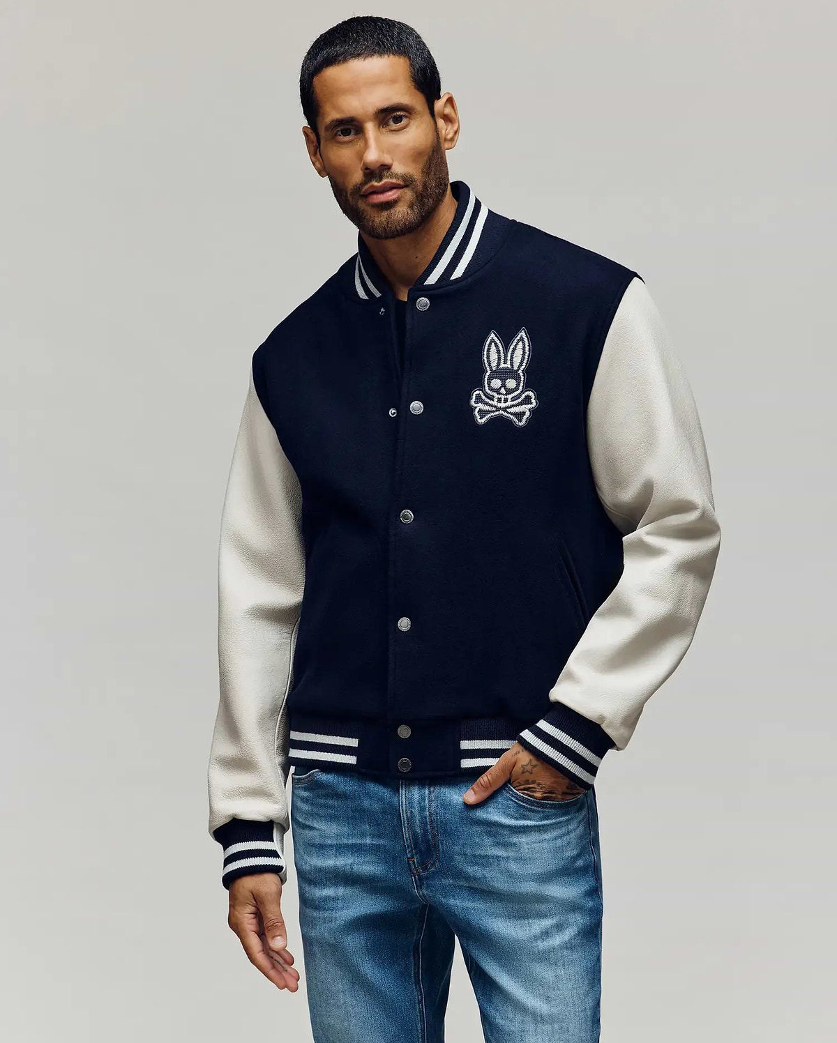 Mens Jacob Leather Bomber, a classic fit navy wool blend jacket with cream leather sleeves, featuring a large embroidered white Psycho Bunny skull logo on the chest and striped ribbed knit trim.