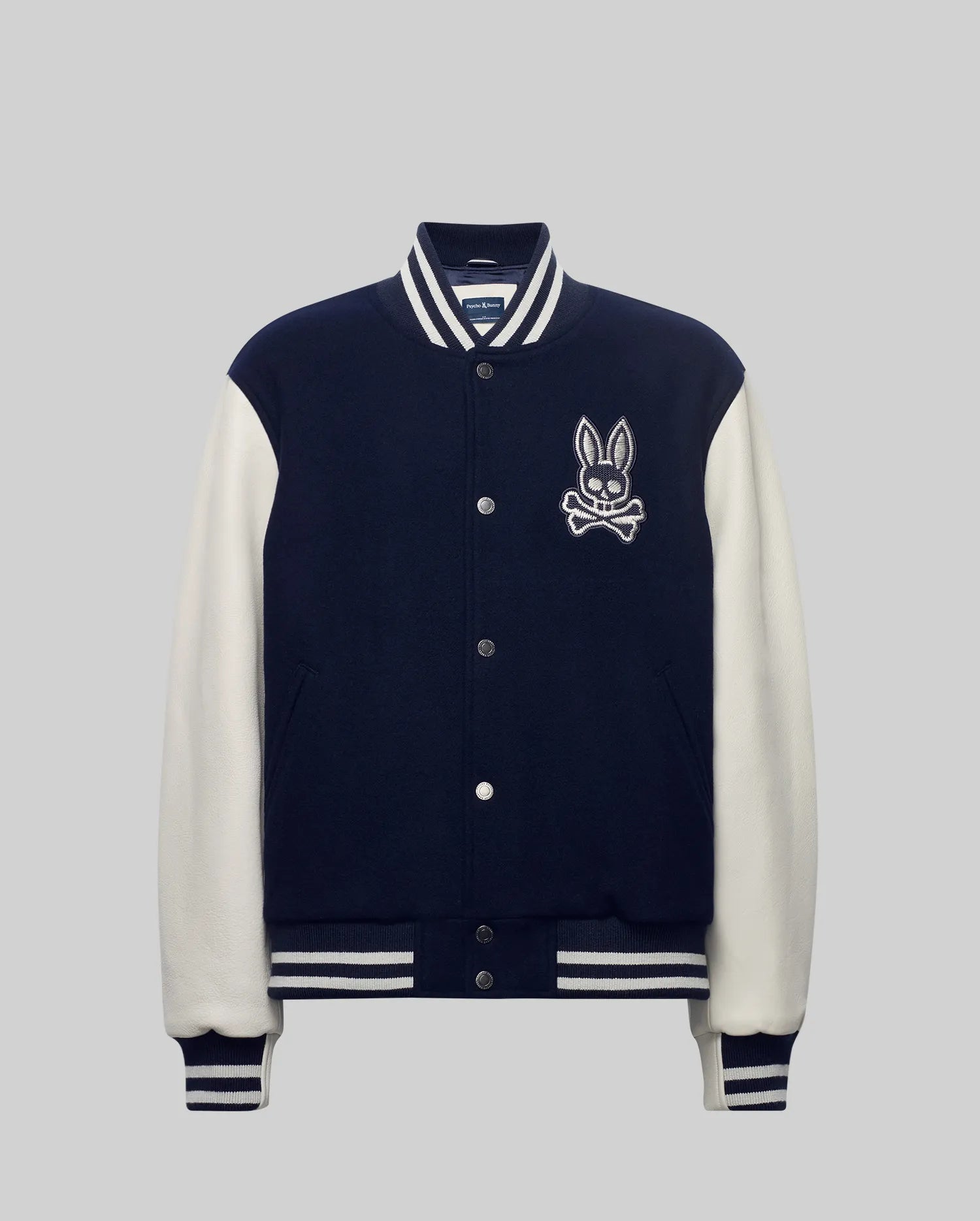 Mens Jacob Leather Bomber in navy with cream leather sleeves, featuring a large embroidered Psycho Bunny skull logo on the left chest, snap buttons, and white striped ribbed knit trim.