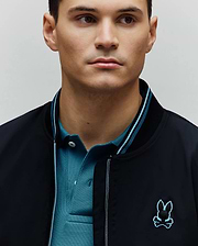 Mens Aldrin Bomber in black, featuring a smooth fabric, ribbed collar with light blue stripes, and an embroidered black and light blue Psycho Bunny skull logo on the chest, layered over a teal pique polo.
