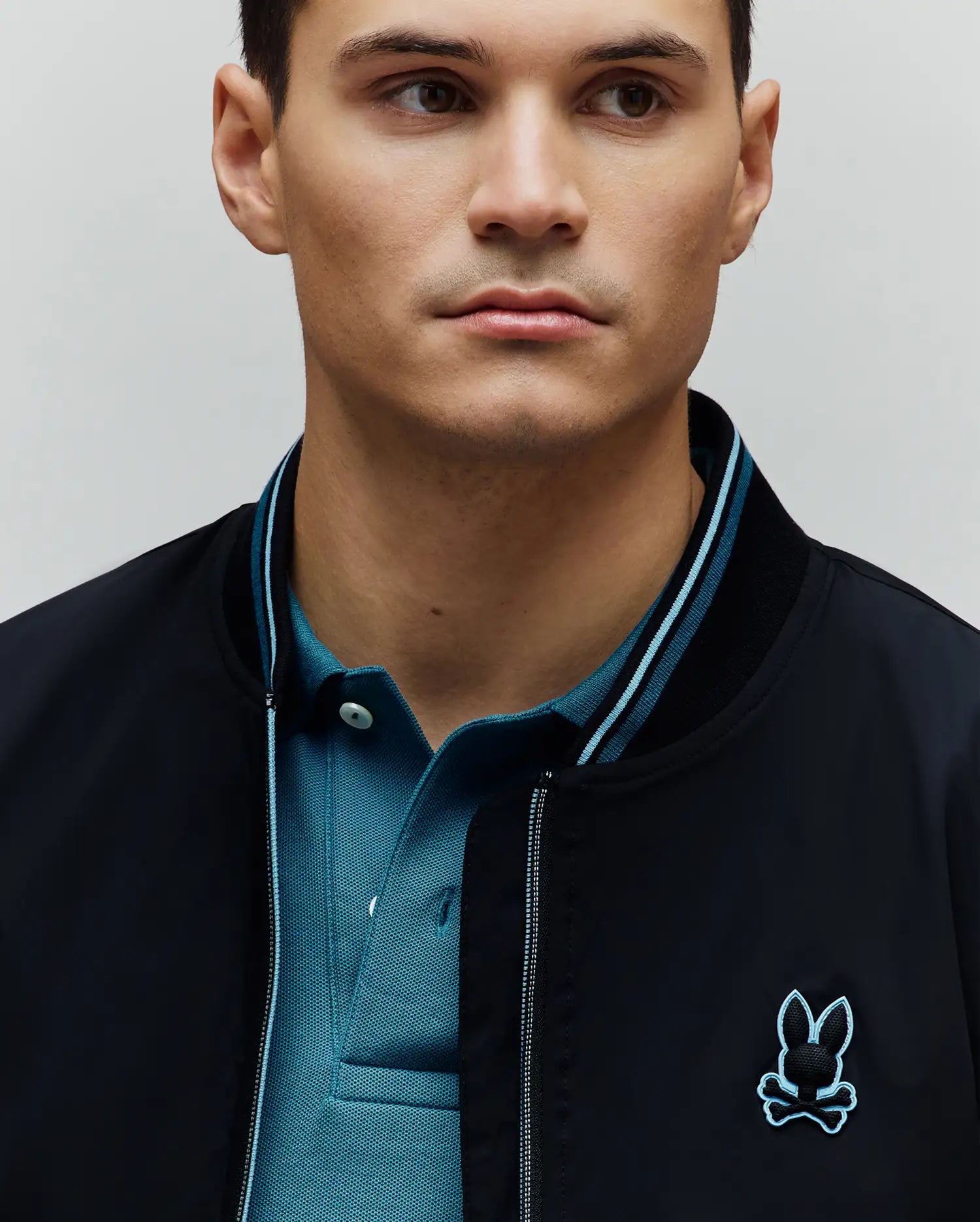 Mens Aldrin Bomber in black, featuring a smooth fabric, ribbed collar with light blue stripes, and an embroidered black and light blue Psycho Bunny skull logo on the chest, layered over a teal pique polo.