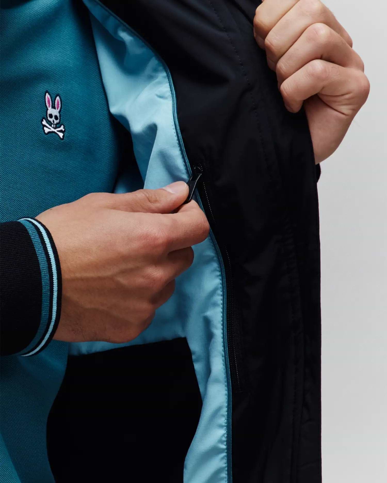 Mens Aldrin Bomber jacket in black with light blue lining, revealing a teal pique polo shirt featuring a small embroidered pink and white Psycho Bunny skull logo on the chest and black ribbed cuffs with light blue stripes.