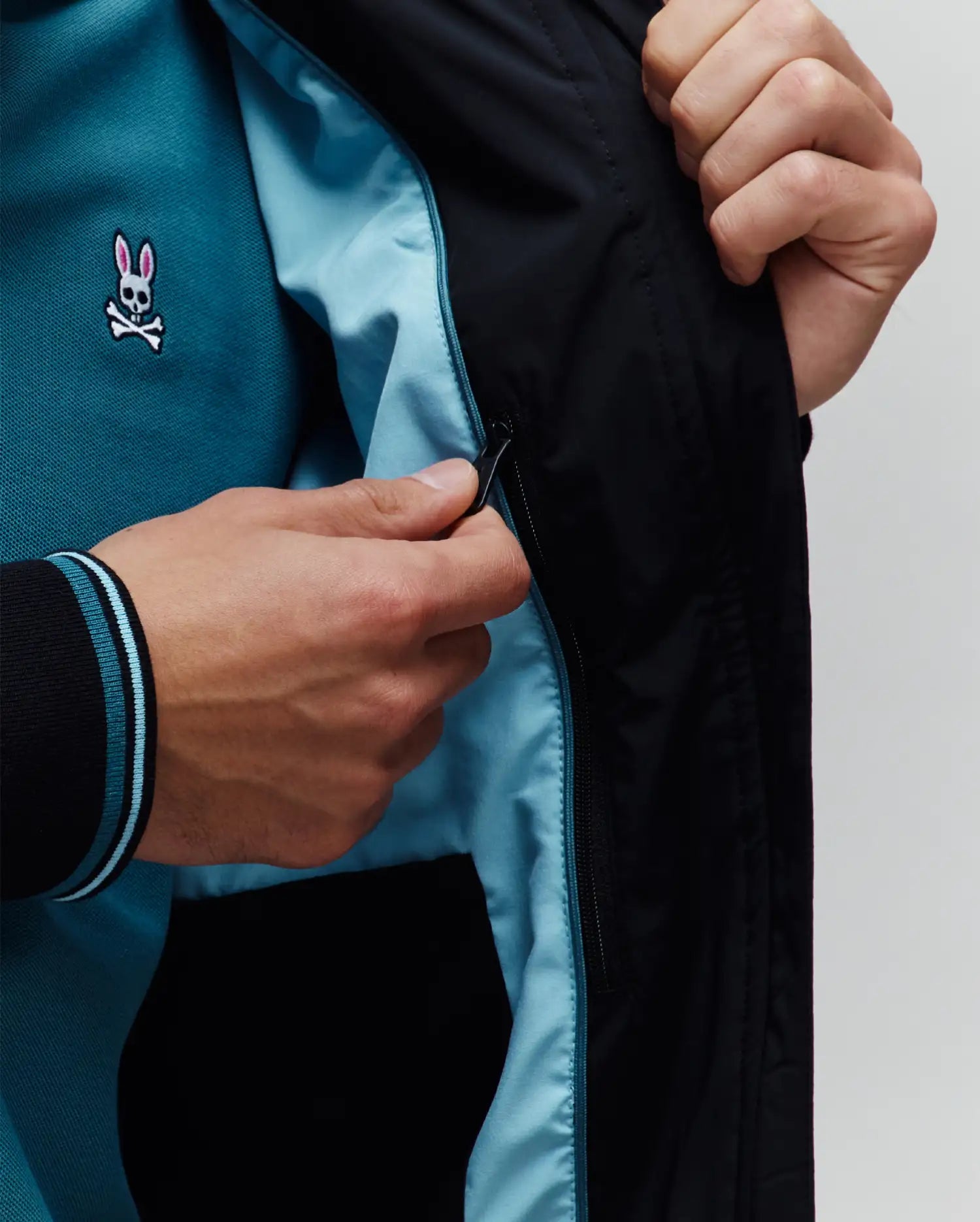 Mens Aldrin Bomber jacket in black with light blue lining, revealing a teal pique polo shirt featuring a small embroidered pink and white Psycho Bunny skull logo on the chest and black ribbed cuffs with light blue stripes.