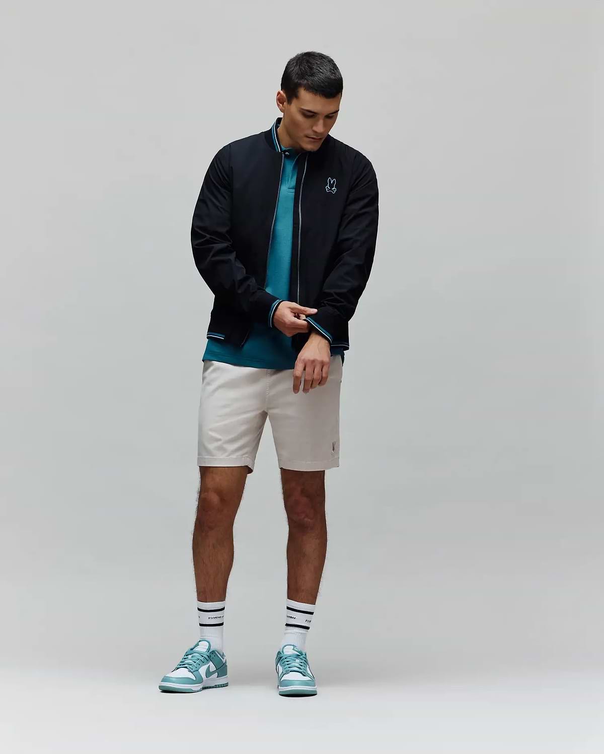 Mens Aldrin Bomber in black with teal trim and embroidered Psycho Bunny skull logo on chest, layered over a teal pique polo, light beige tailored shorts, white striped Psycho Bunny socks, and teal and white sneakers.