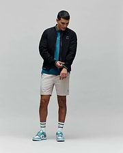 Mens Aldrin Bomber in black with teal trim and embroidered Psycho Bunny skull logo on chest, layered over a teal pique polo, light beige tailored shorts, white striped Psycho Bunny socks, and teal and white sneakers.