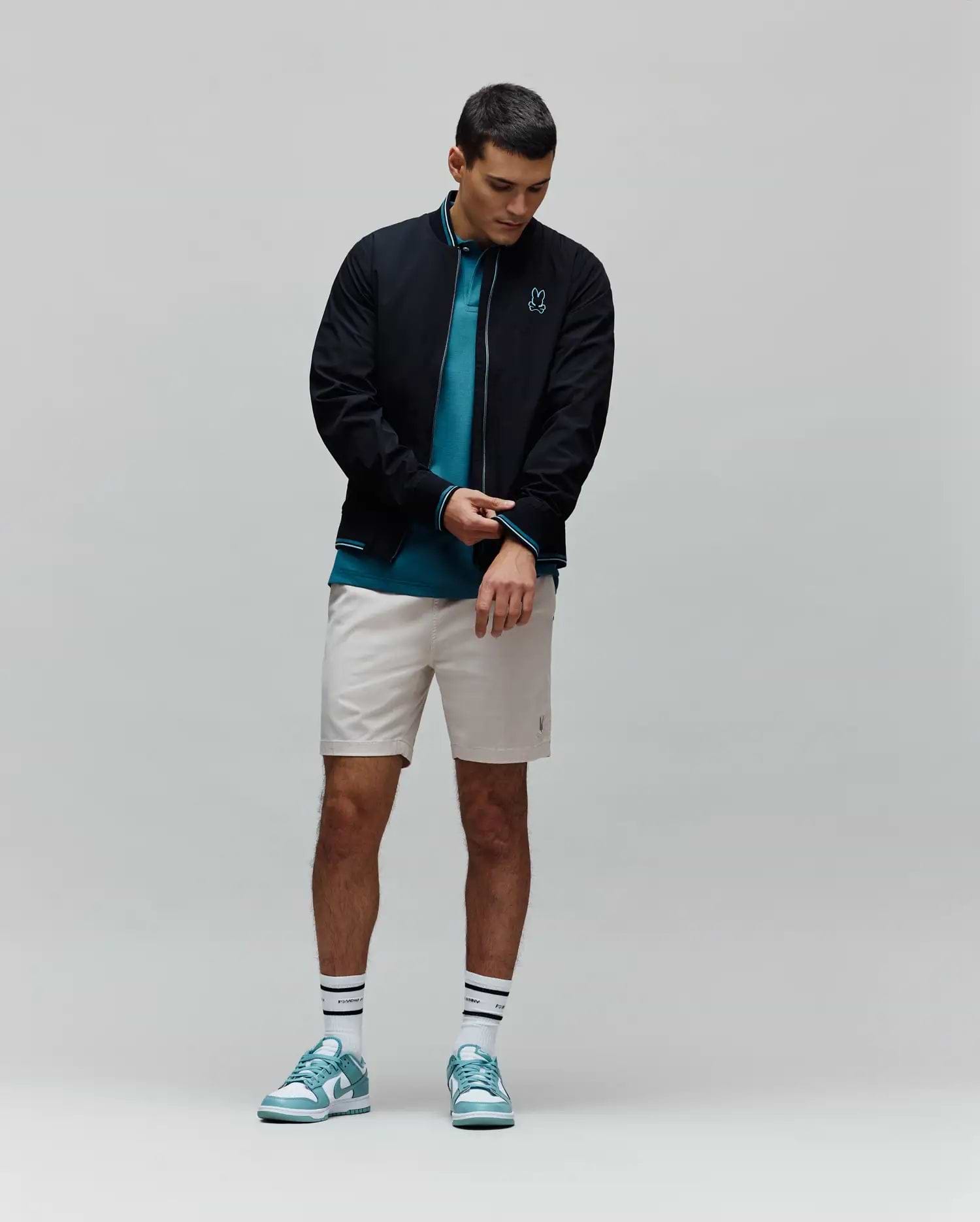 Mens Aldrin Bomber in black with teal trim and embroidered Psycho Bunny skull logo on chest, layered over a teal pique polo, light beige tailored shorts, white striped Psycho Bunny socks, and teal and white sneakers.