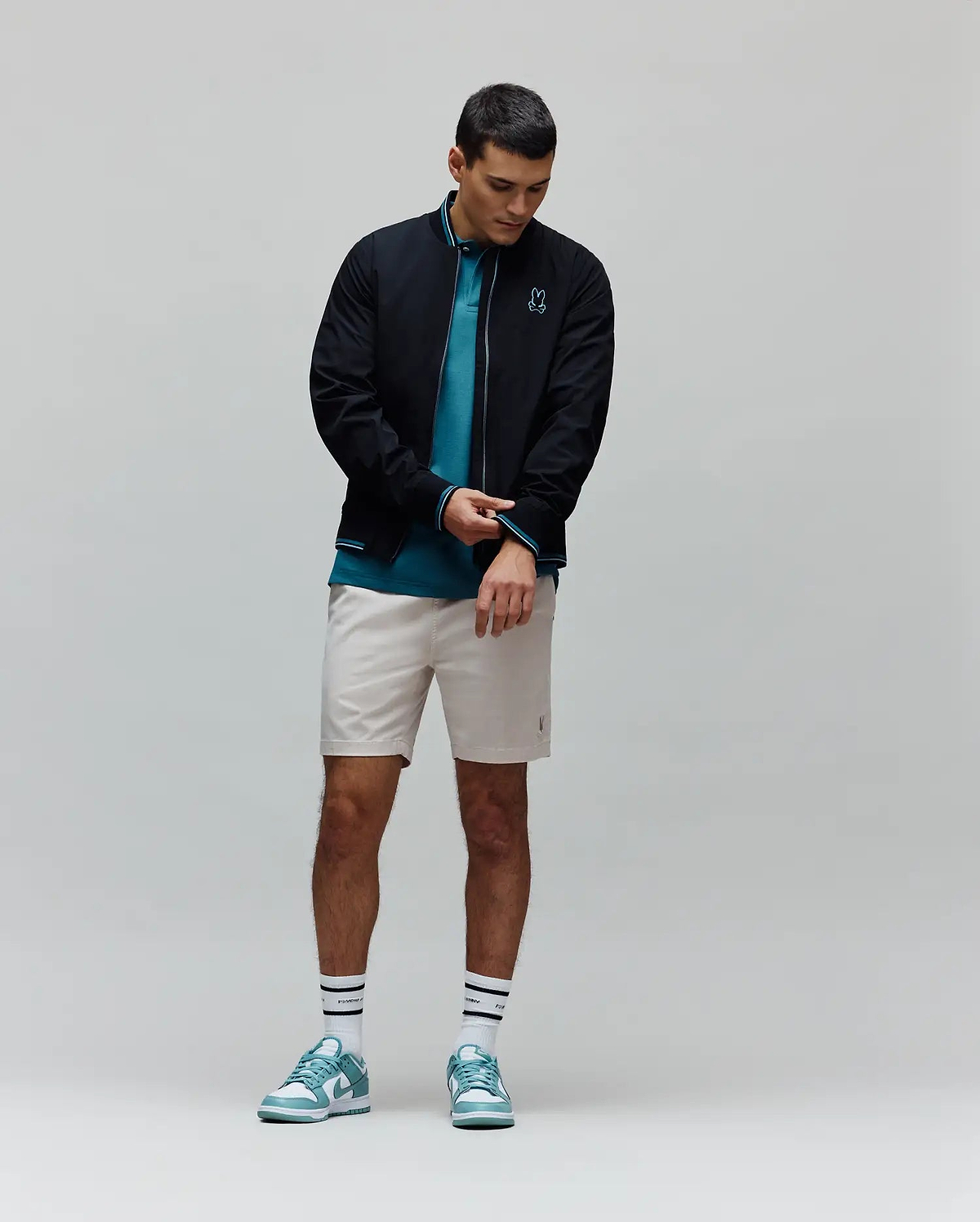 Mens Aldrin Bomber in black with teal trim and embroidered Psycho Bunny skull logo on chest, layered over a teal pique polo, light beige tailored shorts, white striped Psycho Bunny socks, and teal and white sneakers.