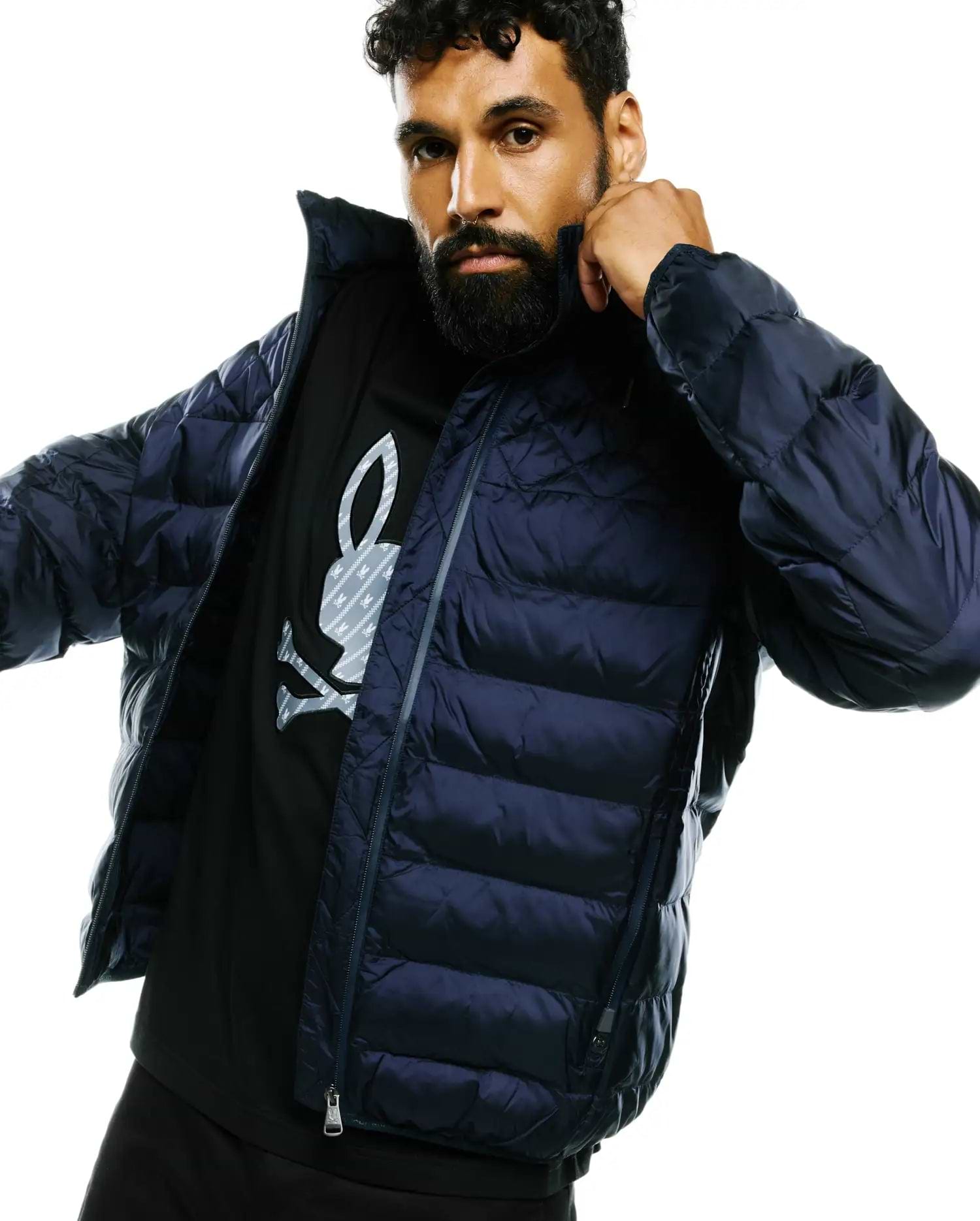 Mens Emerik Hooded Puffer Jacket in dark navy, with horizontal quilting and full zip. Layered over a black jersey t-shirt featuring a large grey Psycho Bunny skull logo with subtle pattern on the chest.