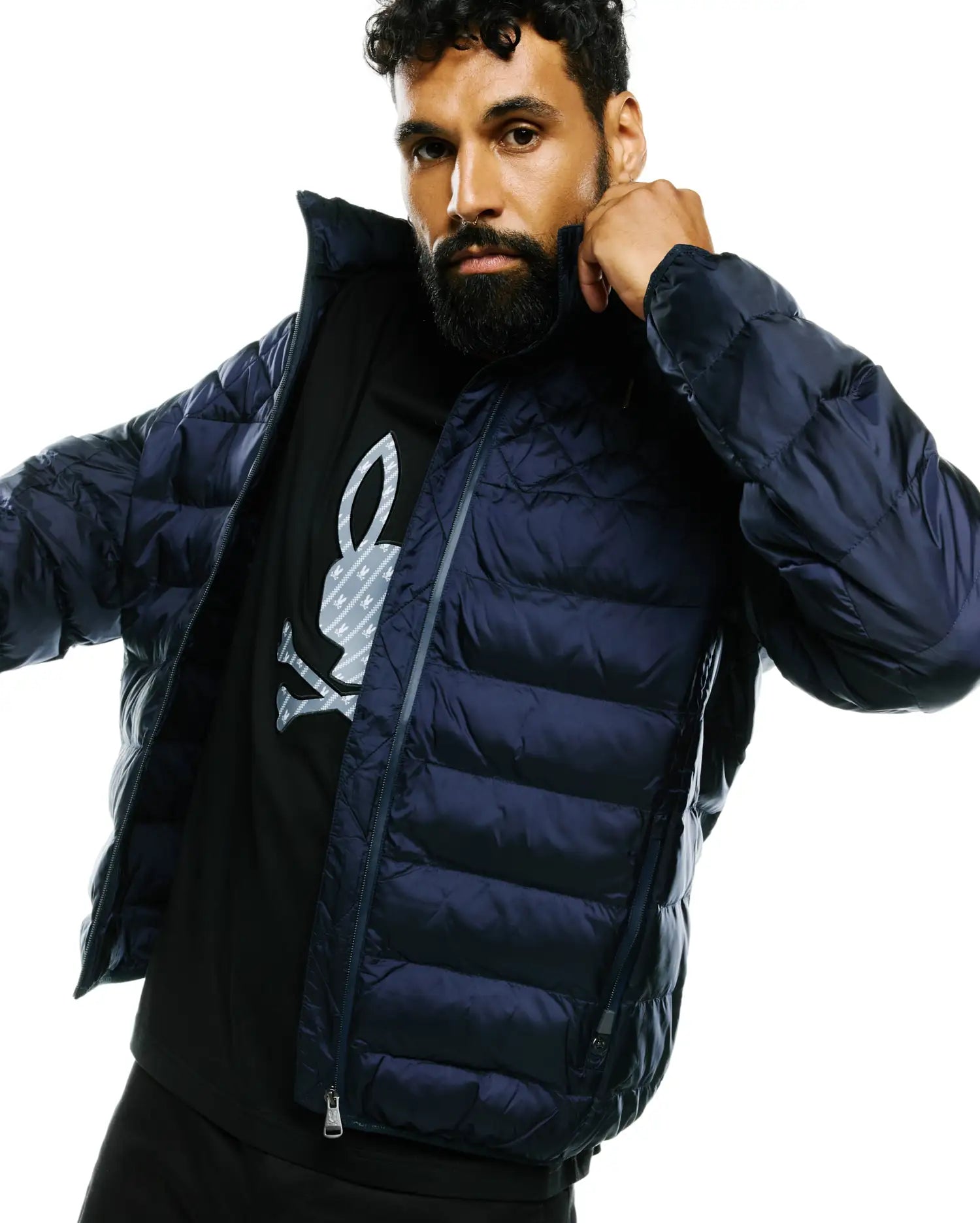 Mens Emerik Hooded Puffer Jacket in dark navy, with horizontal quilting and full zip. Layered over a black jersey t-shirt featuring a large grey Psycho Bunny skull logo with subtle pattern on the chest.