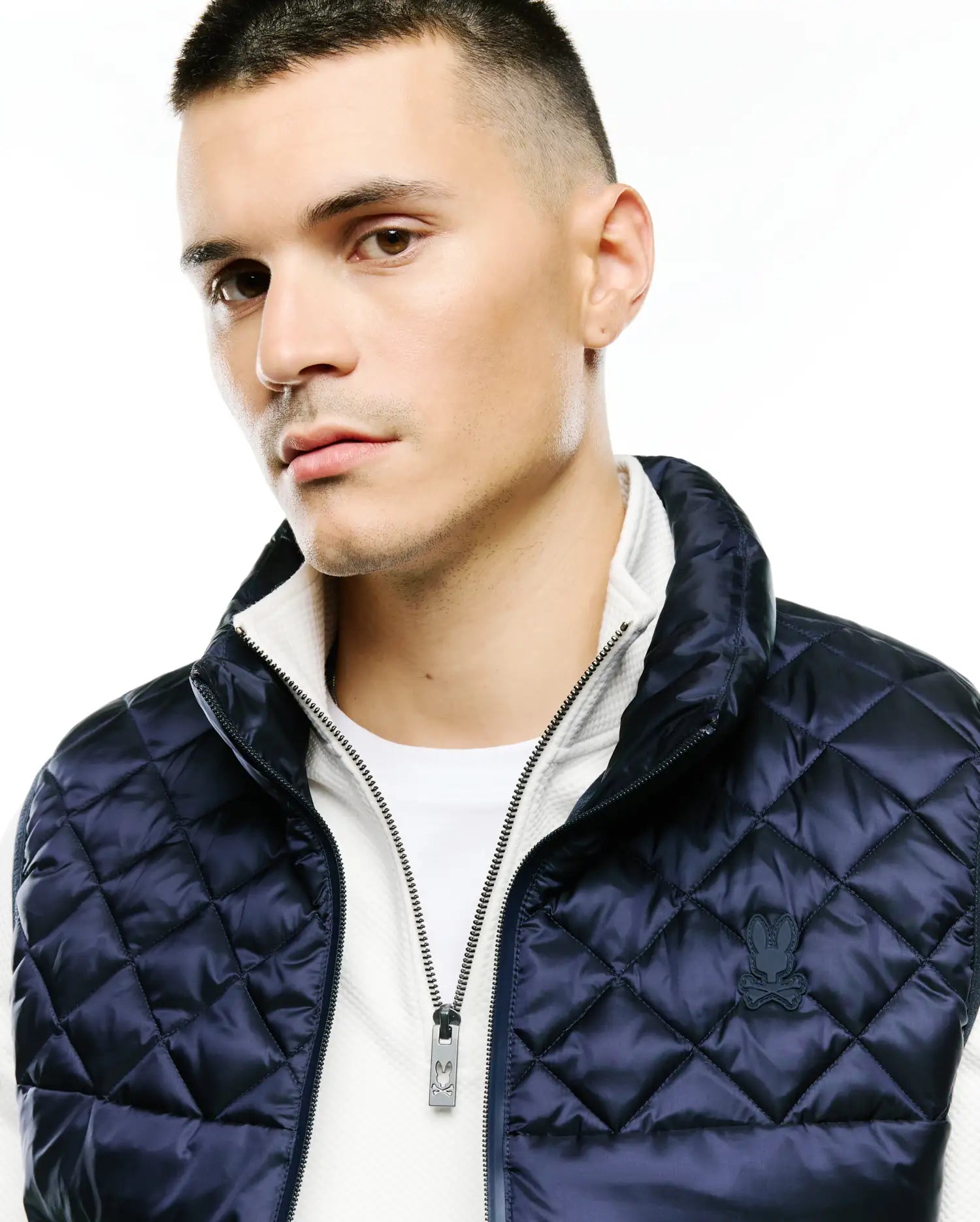 Mens Emerik Quilted Puffer Vest in navy, diamond quilt pattern, white stand collar, full zip with silver Psycho Bunny logo pull, and embroidered bunny skull on left chest.