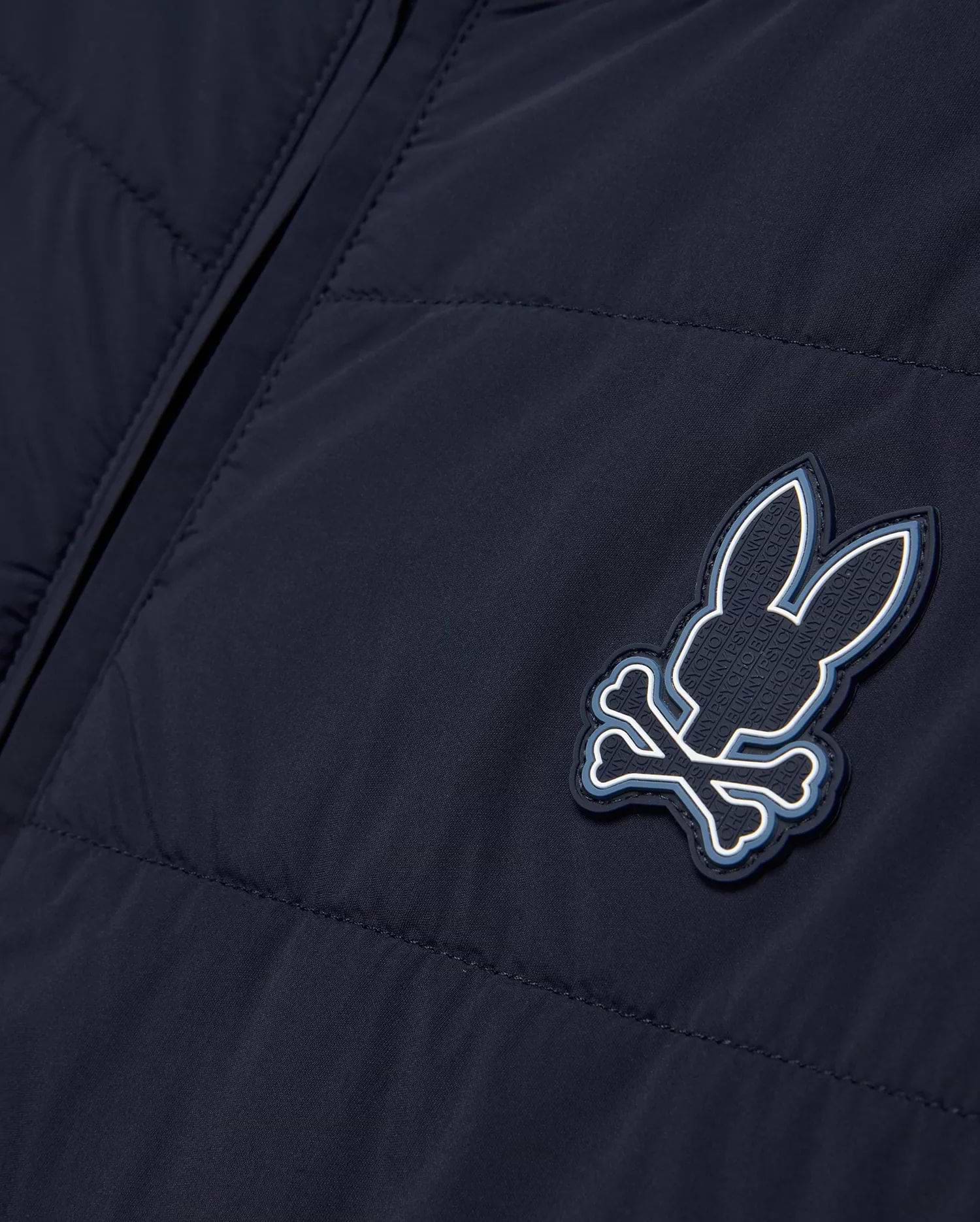 Mens Gordley Mix Media Jacket in navy blue, featuring a quilted left panel and smooth right panel, with a prominent Psycho Bunny skull and crossbones logo on the chest, detailed with white and light blue accents and embossed brand lettering. (5)