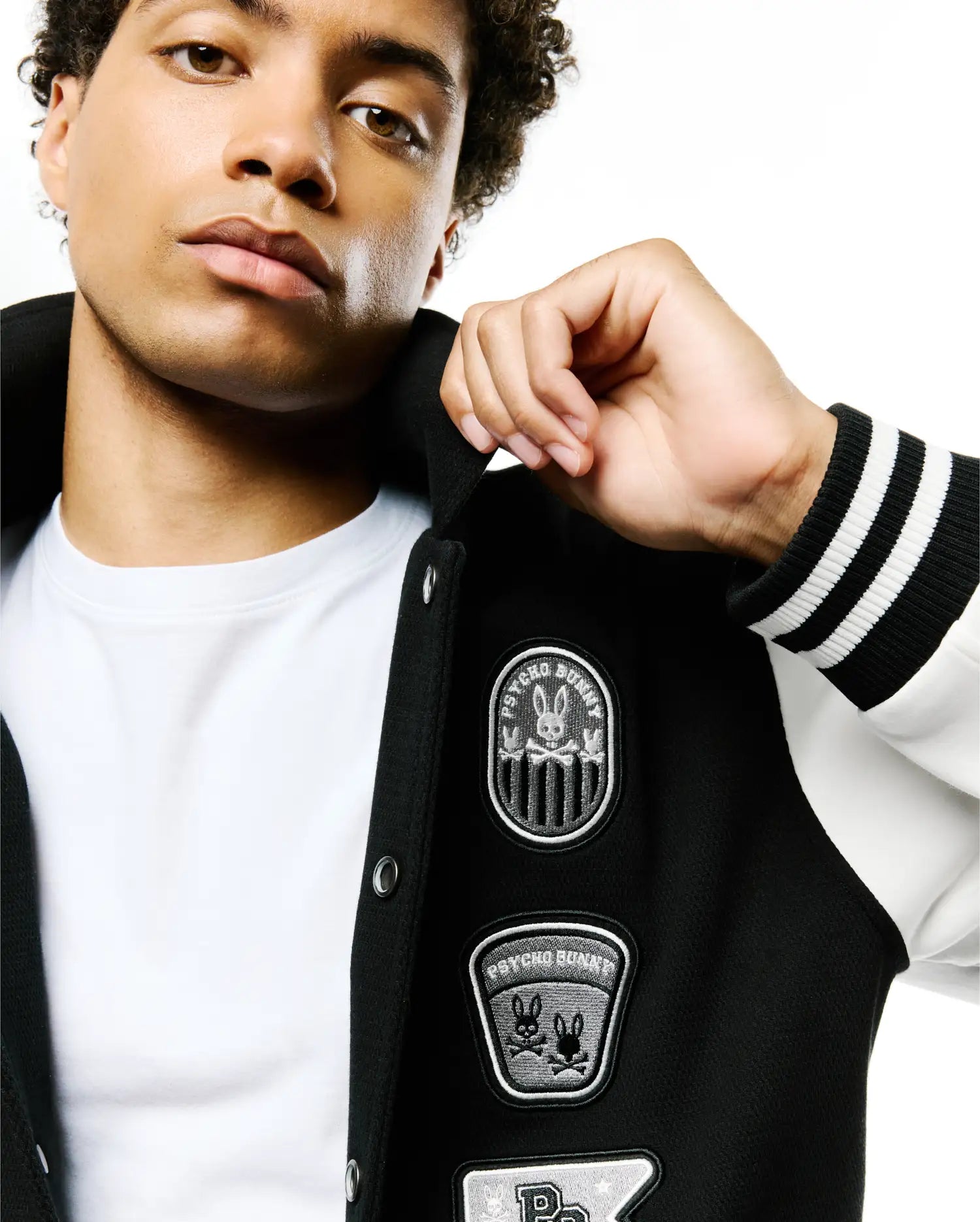 Mens Reilly Knit Coach Jacket, black with white sleeves, features multiple embroidered Psycho Bunny skull patches on the chest, ribbed cuffs with white stripes, and a classic fit. (3)