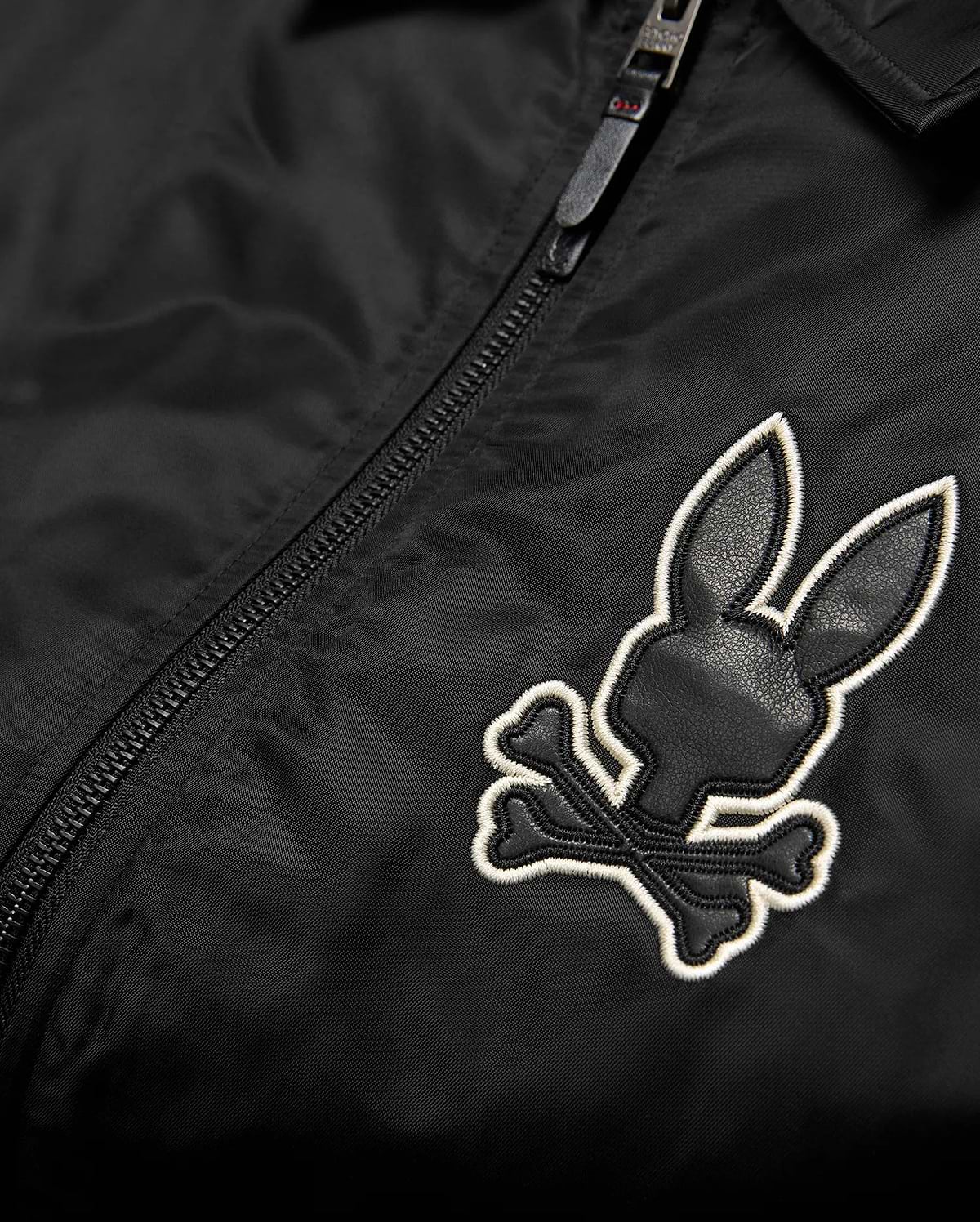 Mens Nathaniel Satin Coach Jacket in black, featuring a prominent embroidered Psycho Bunny skull logo on the chest with black leather-like texture and a white outline, alongside a black zipper. (6)