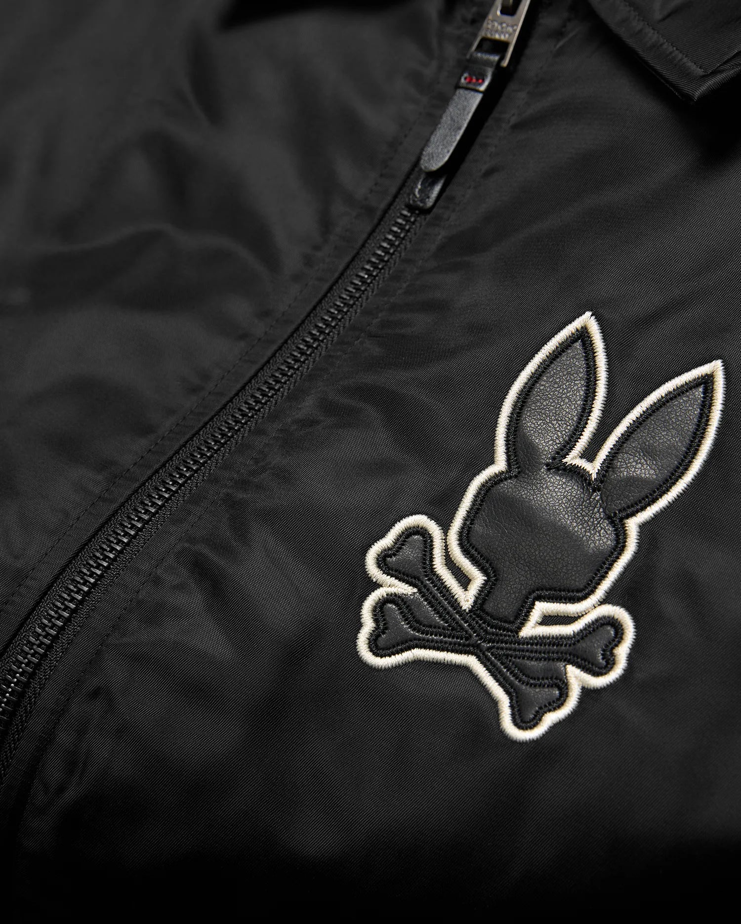 Mens Nathaniel Satin Coach Jacket in black, featuring a prominent embroidered Psycho Bunny skull logo on the chest with black leather-like texture and a white outline, alongside a black zipper. (6)