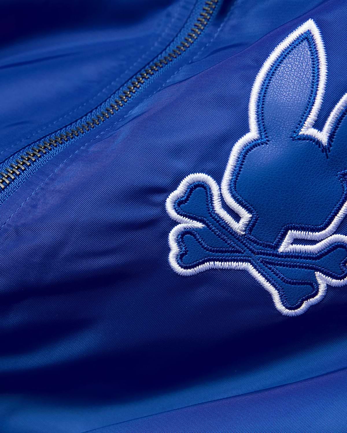 Mens Nathaniel Satin Coach Jacket in royal blue satin, featuring a large, embroidered Psycho Bunny skull logo with a dark blue textured fill and thick white outline, alongside a silver zipper. (7)