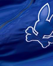 Mens Nathaniel Satin Coach Jacket in royal blue satin, featuring a large, embroidered Psycho Bunny skull logo with a dark blue textured fill and thick white outline, alongside a silver zipper. (7)