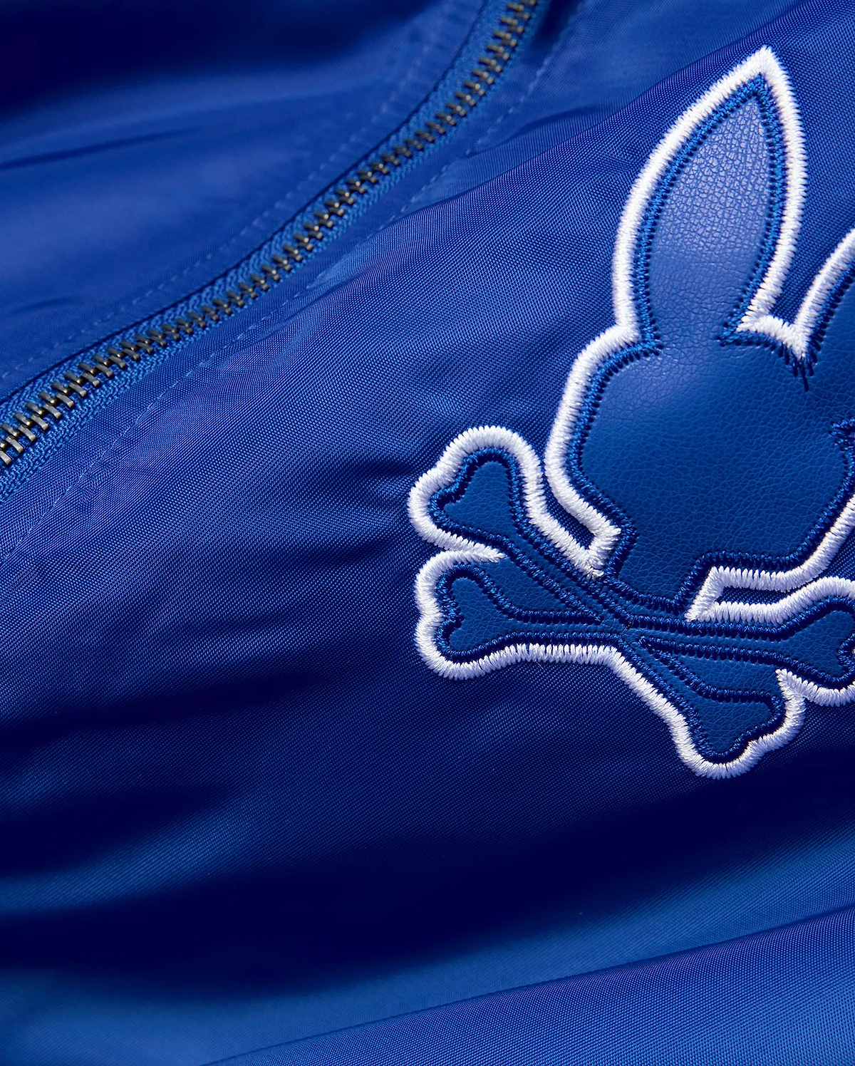 Mens Nathaniel Satin Coach Jacket in royal blue satin, featuring a large, embroidered Psycho Bunny skull logo with a dark blue textured fill and thick white outline, alongside a silver zipper. (7)
