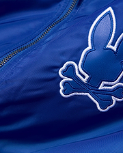 Mens Nathaniel Satin Coach Jacket in royal blue satin, featuring a large, embroidered Psycho Bunny skull logo with a dark blue textured fill and thick white outline, alongside a silver zipper. (7)