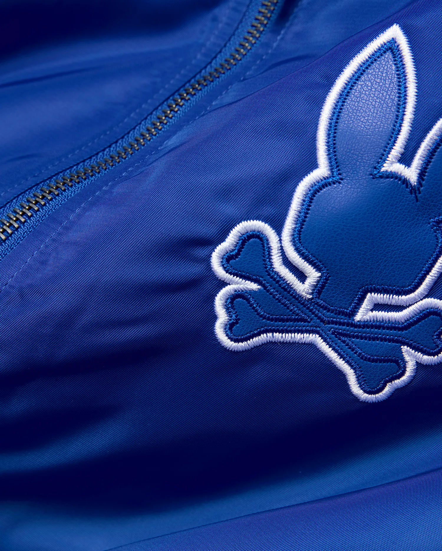 Mens Nathaniel Satin Coach Jacket in royal blue satin, featuring a large, embroidered Psycho Bunny skull logo with a dark blue textured fill and thick white outline, alongside a silver zipper. (7)