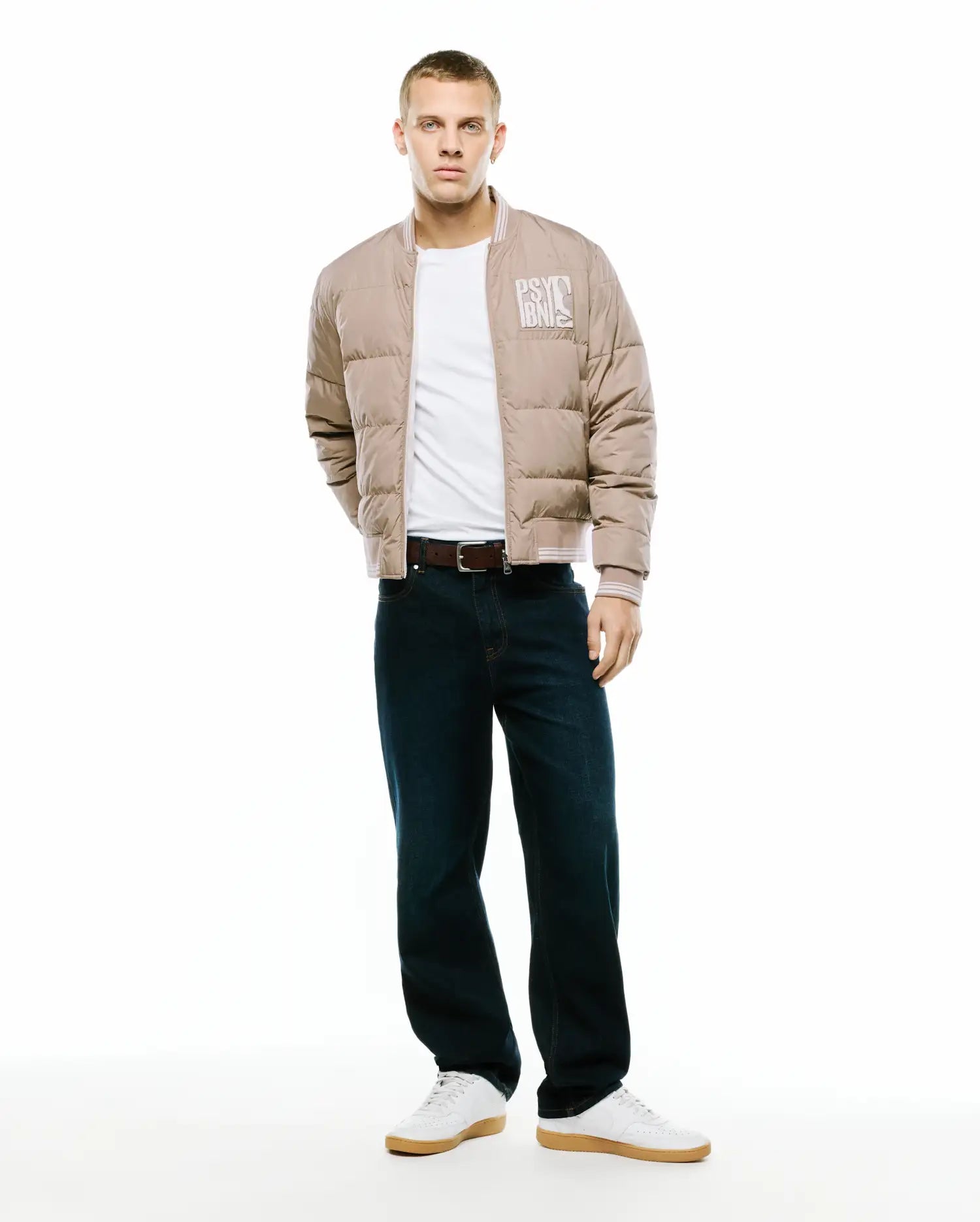 Men's Franco Reversible Bomber: Tan padded bomber jacket with white ribbed collar, cuffs, and hem, featuring a large white Psycho Bunny text logo on the left chest. (1)