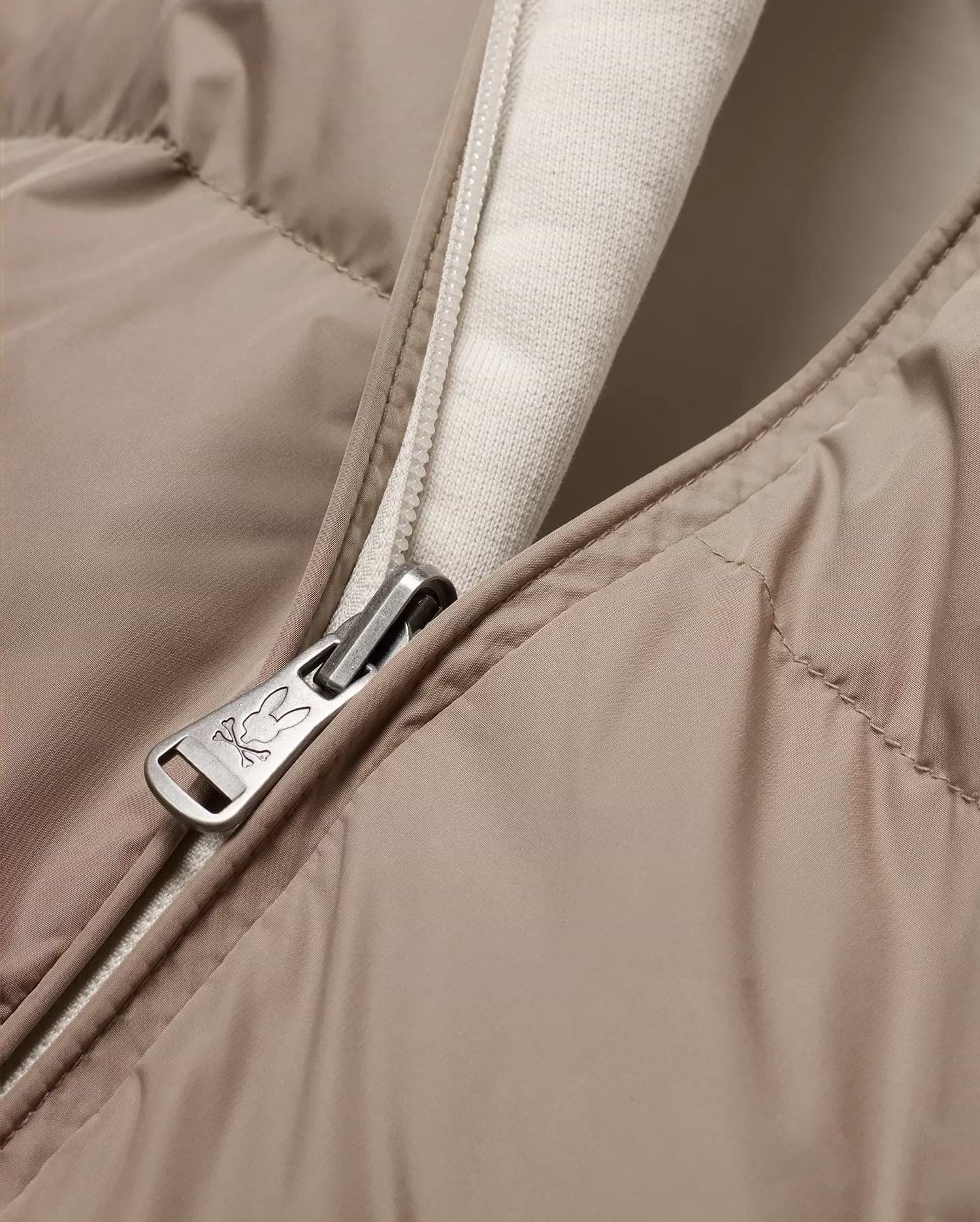 Men's Franco Reversible Bomber, a beige padded jacket with a cream knit lining and a full-zip closure featuring a silver Psycho Bunny skull logo engraved zipper pull. (6)