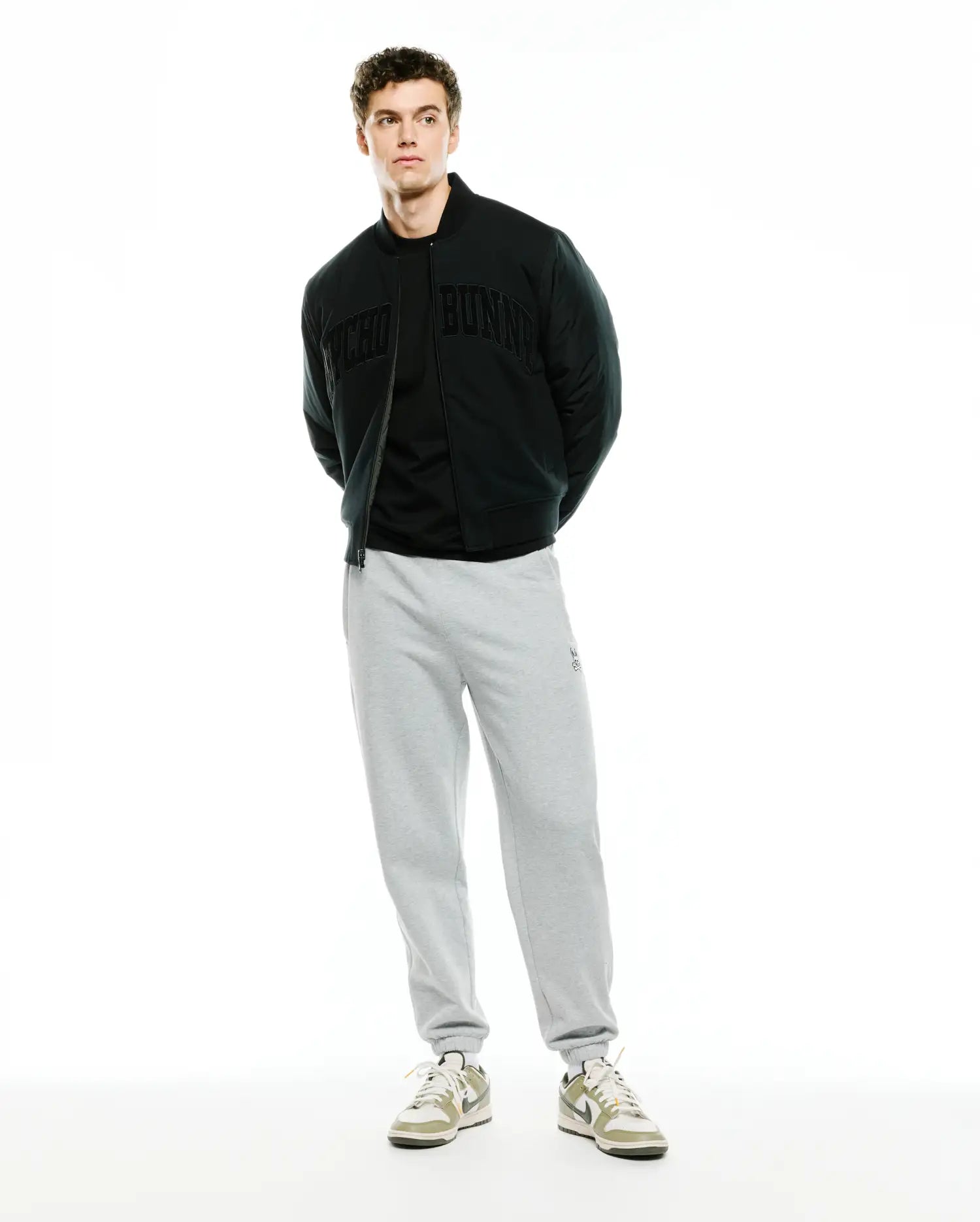 Mens Joaquin Mix Media Reversible Bomber in black, featuring large tonal Psycho Bunny lettering across the chest, paired with light grey relaxed fit french terry sweatpants with a small embroidered bunny skull logo on the left thigh.
