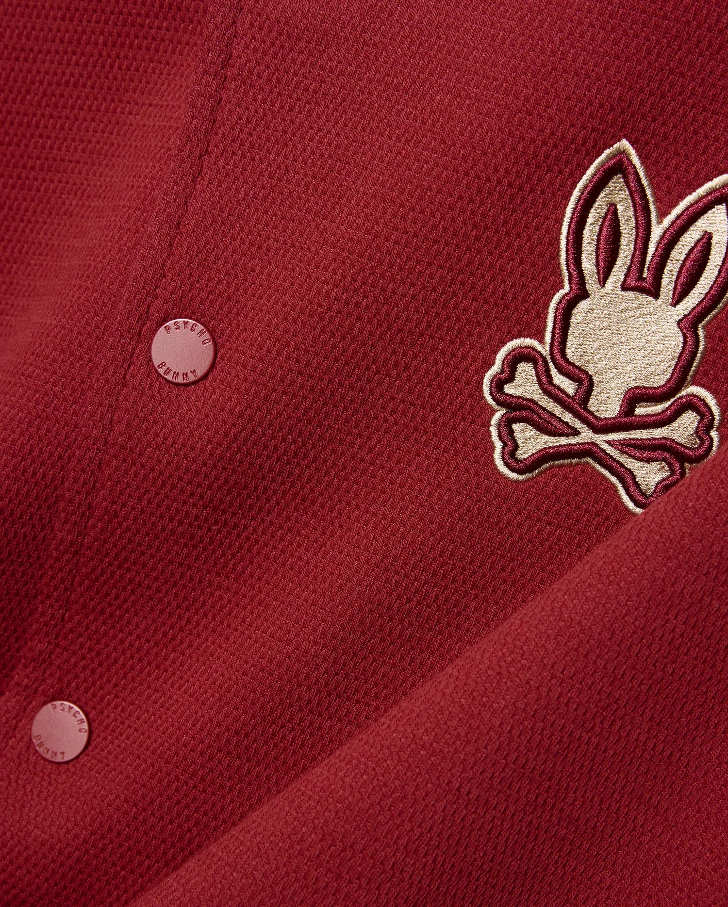 Mens Reilly Knit Bomber in deep red pique knit, featuring a large embroidered gold and burgundy Psycho Bunny skull logo on the chest and matching branded buttons.