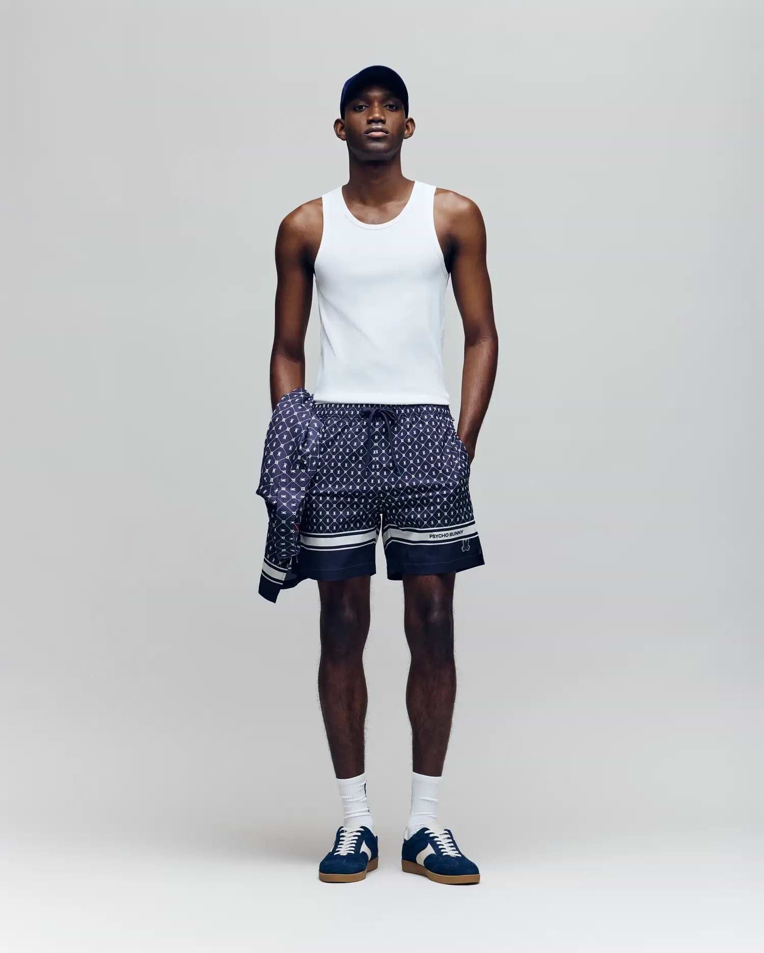 A man stands against a plain background wearing a Psycho Bunny MENS 2 PACK TANK TOP - MO1400001 in white, patterned navy shorts, a matching long-sleeve shirt tied at his waist, a navy cap, white socks, and navy sneakers.