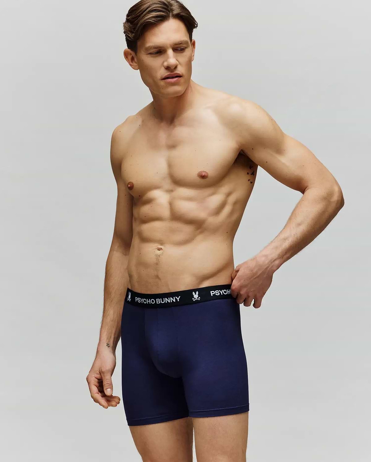 A muscular man stands against a plain background wearing Psycho Bunny MEN'S 3 PACK BOXER BRIEF (MO1400006) in navy blue, featuring a black waistband with “Psycho Bunny” in white text. He looks to the side, one hand touching the waistband.